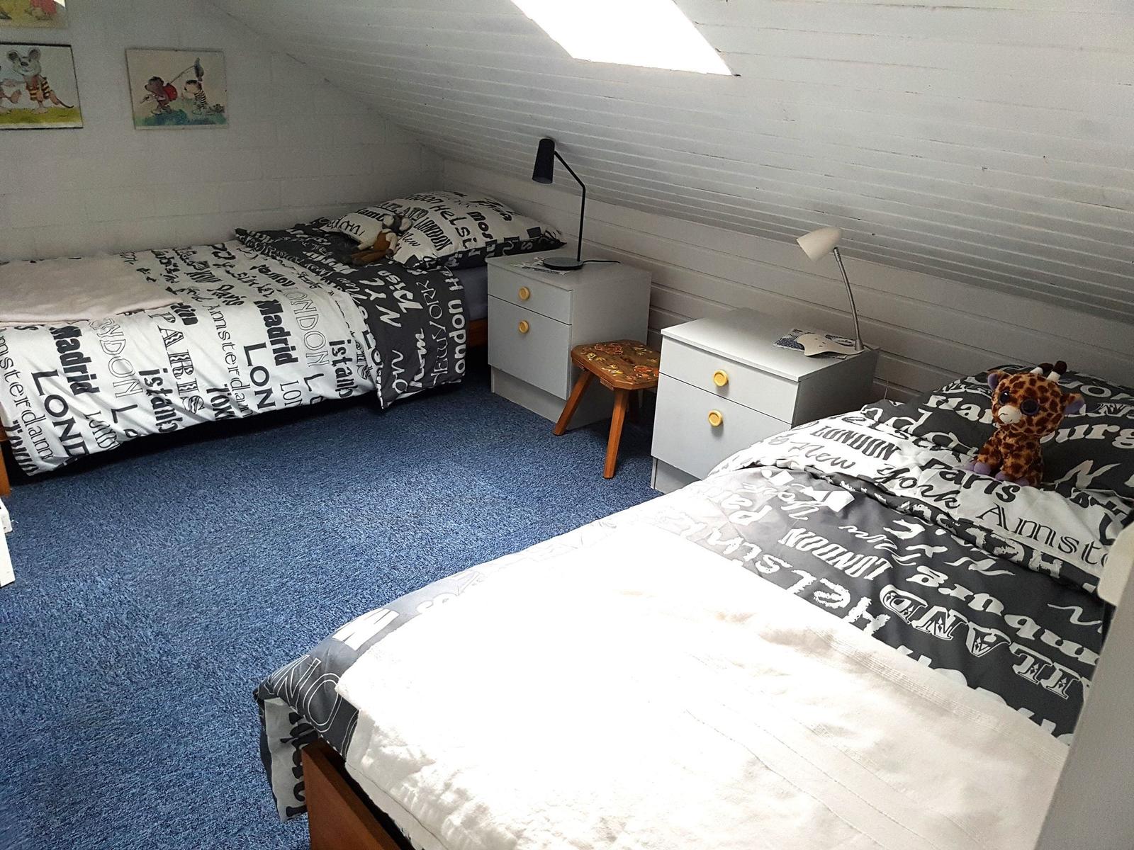 Zwei Betten mit Stoffbezug, Nachttische und Decken mit Stadtbezeichnungen.