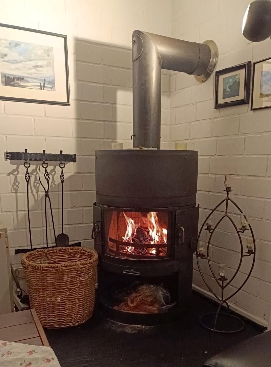 Kaminofen mit brennendem Feuer, Kaminrohr, Korb und Kerzenhalter an weißer Ziegelwand.