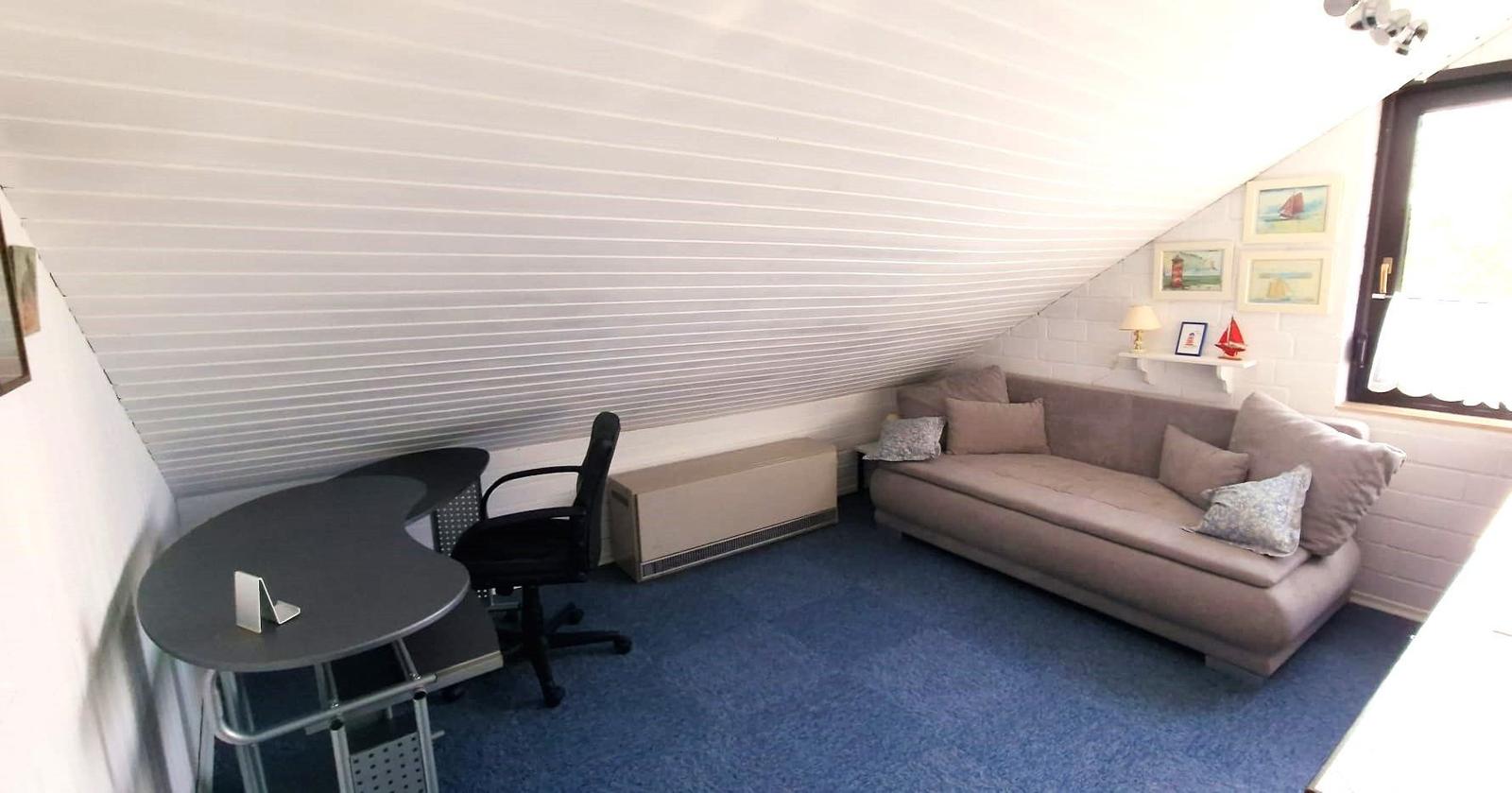 Ein Raum mit Sofa, Schreibtisch und schrägem Dach. Blauer Teppich und Wandbilder.