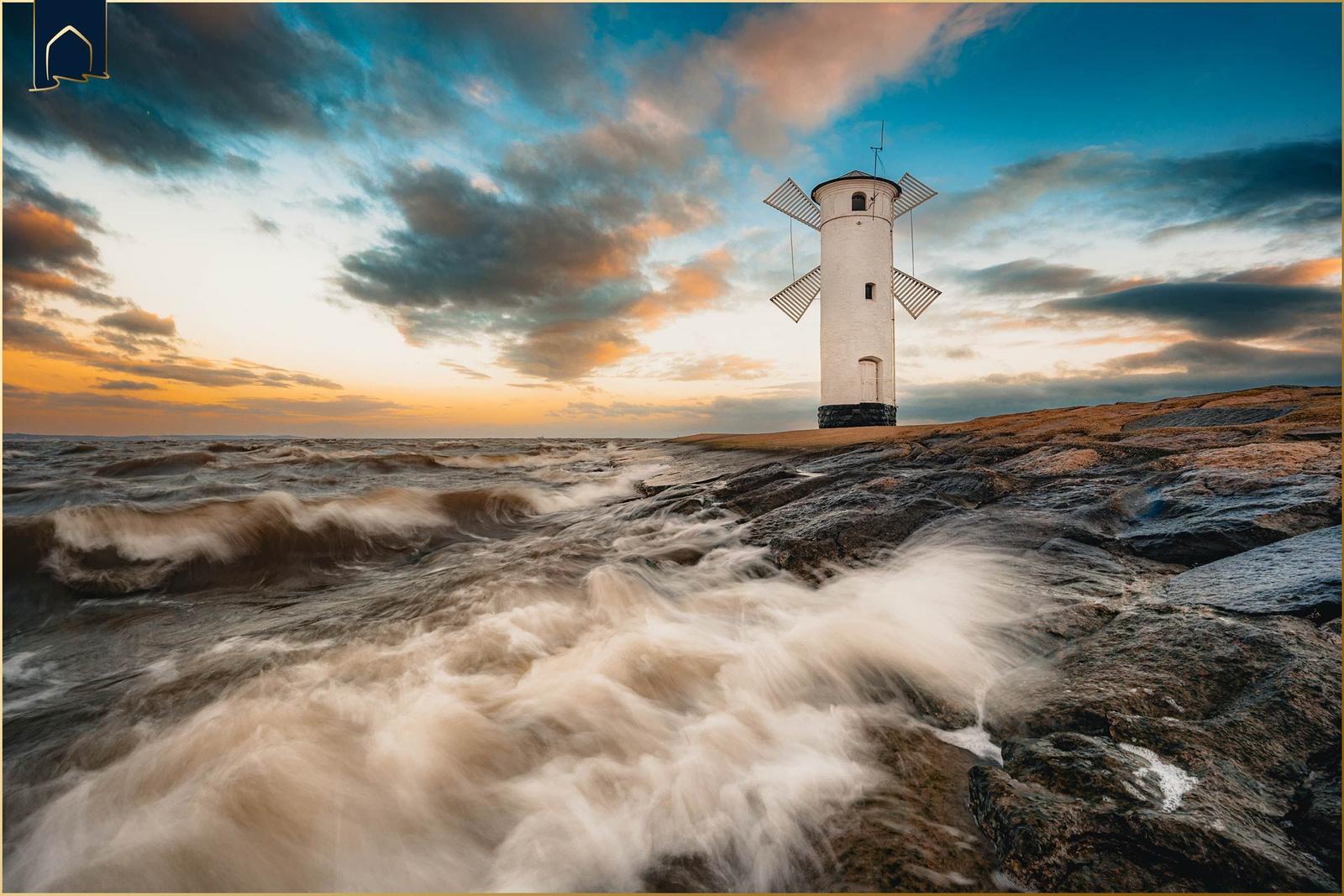 Weißer Windmühlenbau auf felsigem Ufer bei Sonnenuntergang mit welligem Meer.