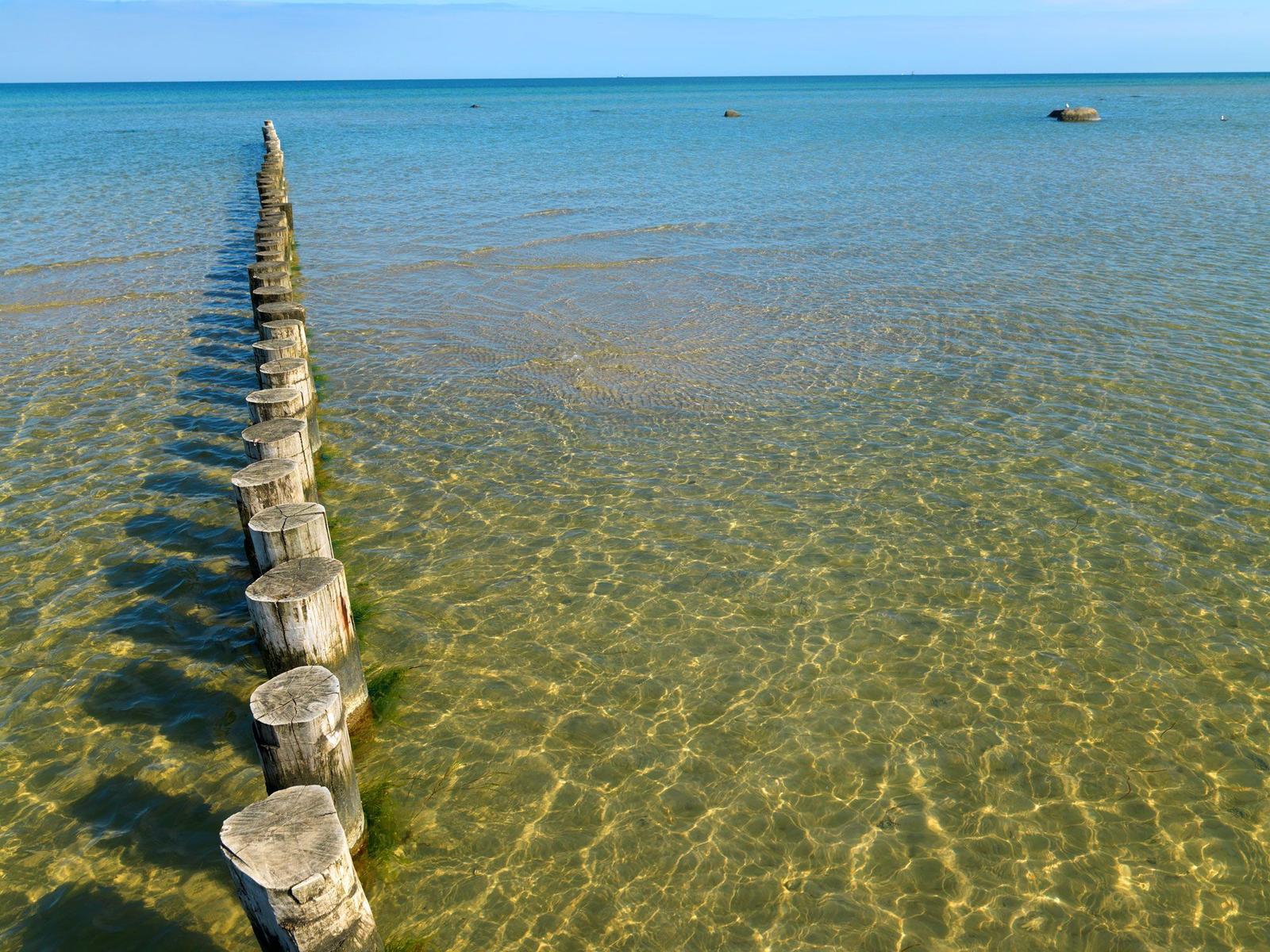 Holzpfähle führen ins klare Meer mit Blick auf fernes Wasser.