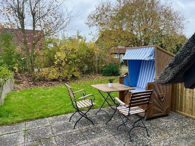 Terrasse mit Tisch, zwei Stühlen und Strandkorb. Grünfläche und Bäume im Hintergrund.