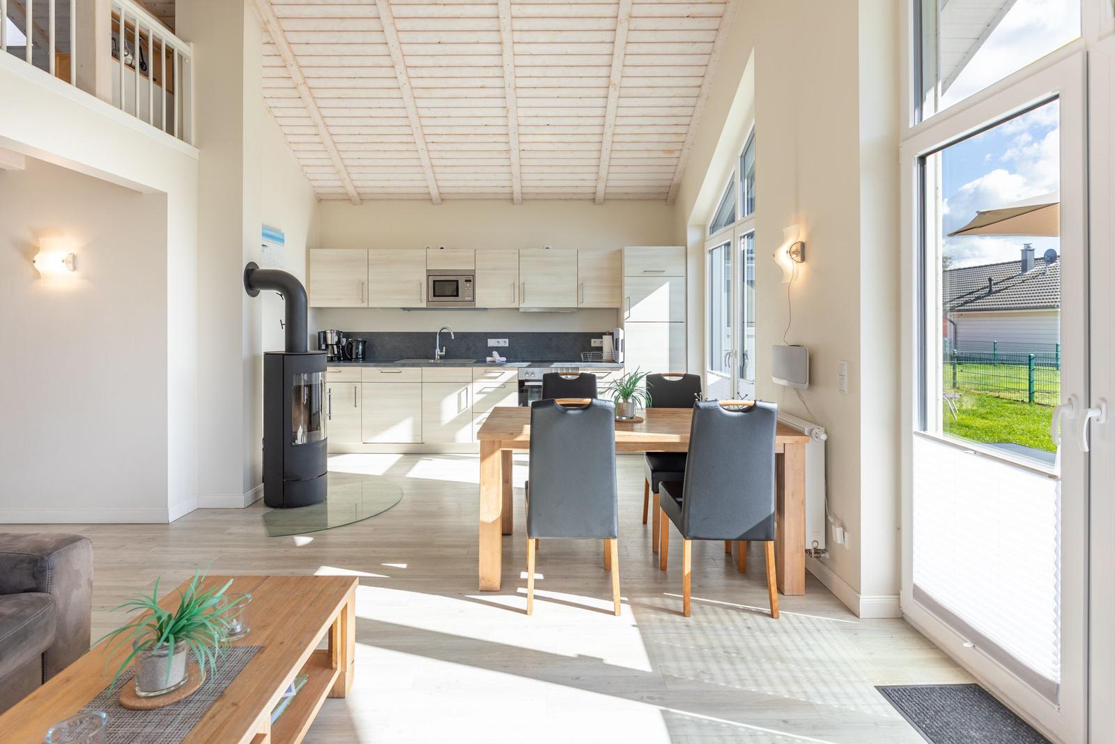 Offener Raum mit Küche, Esstisch und Kaminofen. Große Fenster und Terrasse.