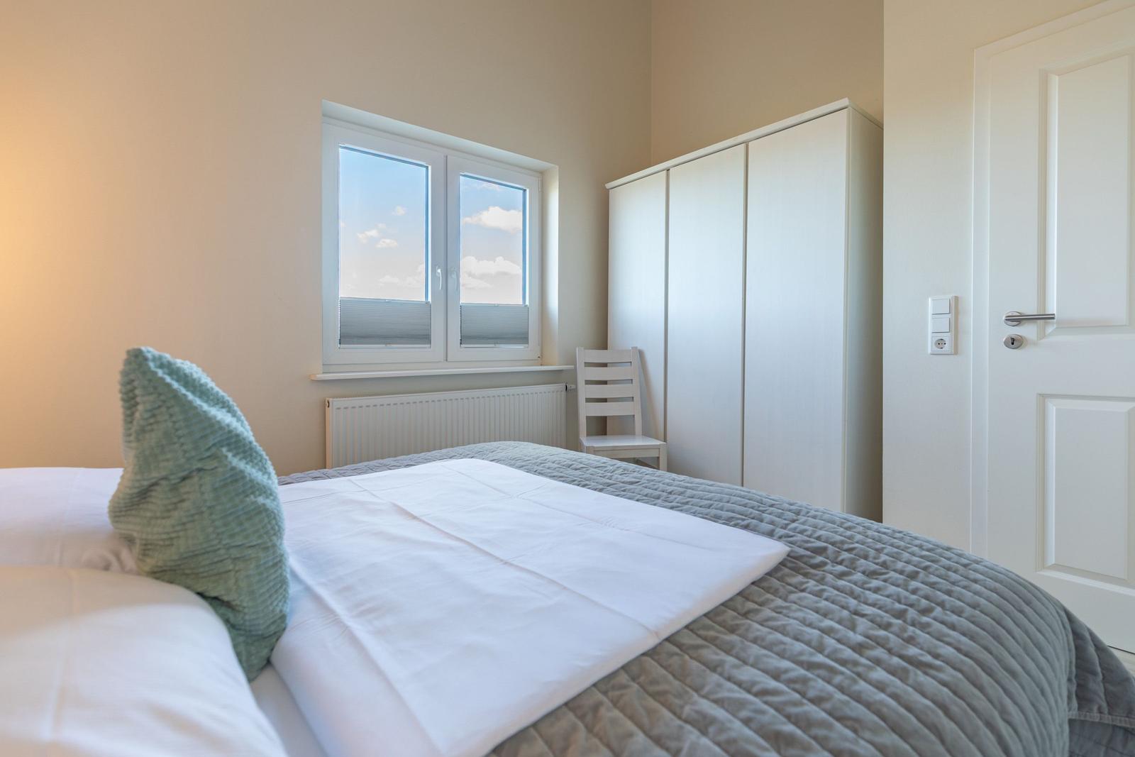 Schlafzimmer mit Bett, Kleiderschrank und Fenster. Tür und Stuhl sind sichtbar.