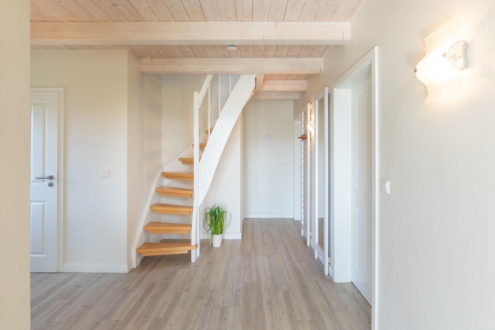 Hausflur mit Holztreppe, Holzboden und Deckenbalken. An der Wand hängt eine Lampe.