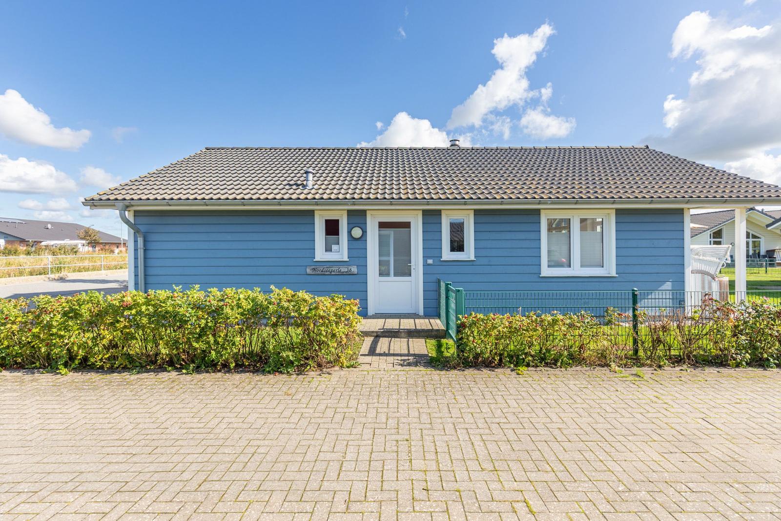 Blau gestrichenes Ferienhaus mit weißen Fenstern und Eingangstür. Vorgarten mit Hecke und gepflastertem Hof.