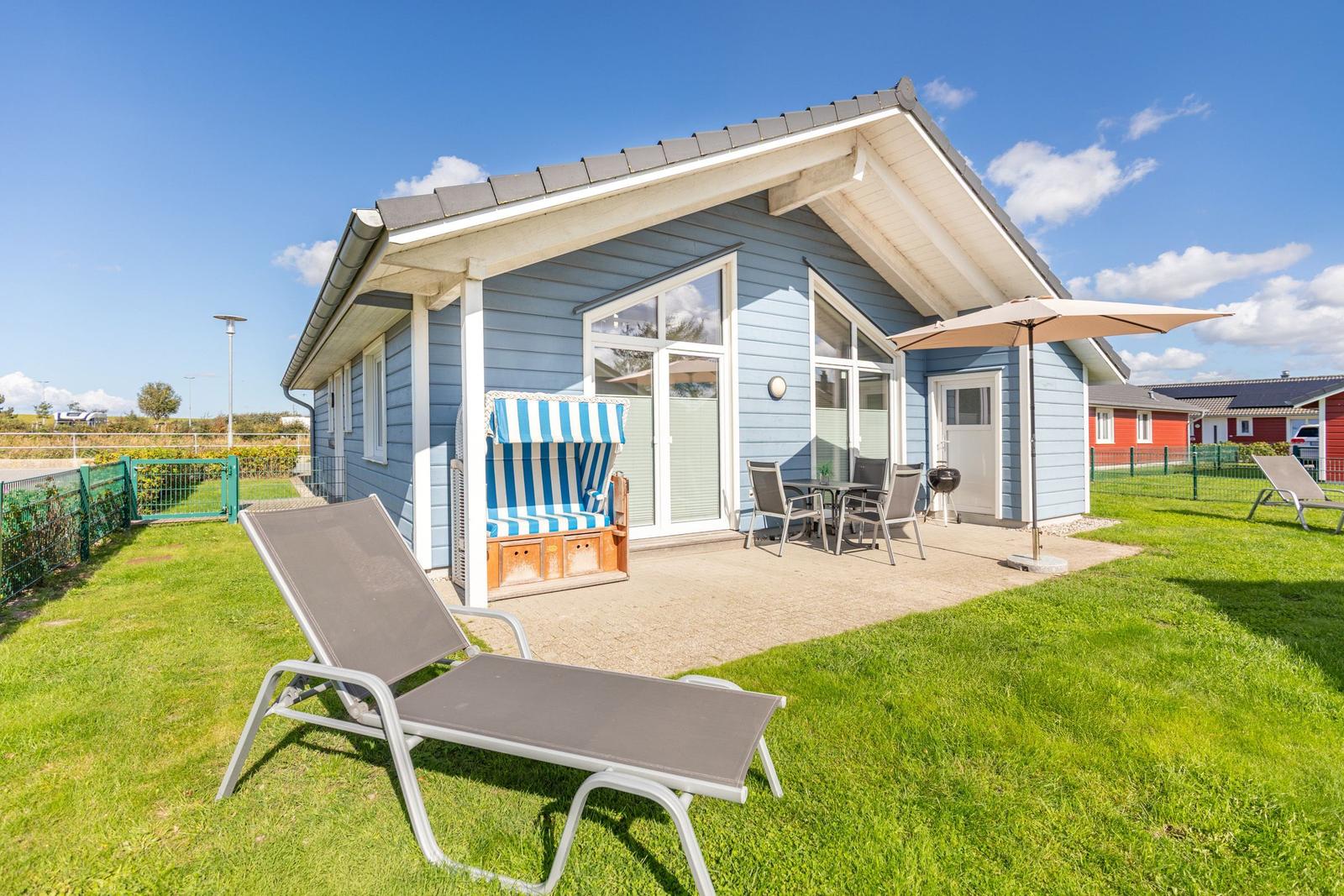 Blauer Ferienhaus mit Terrasse, Liege und Sonnenschirm im Garten.