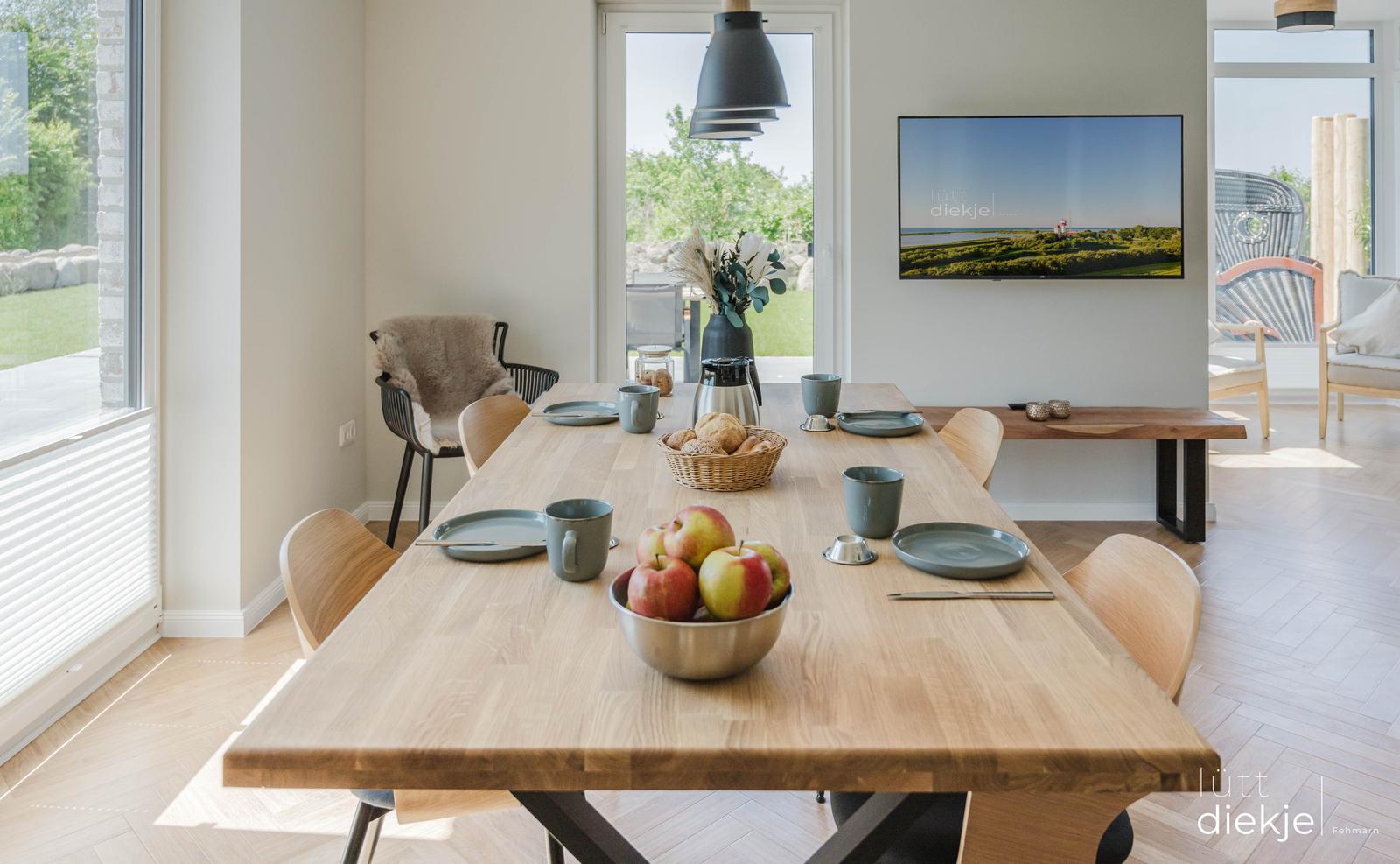 Essbereich mit Holztisch, Stühlen und Obst. Fernseher an der Wand. Blick auf Garten.