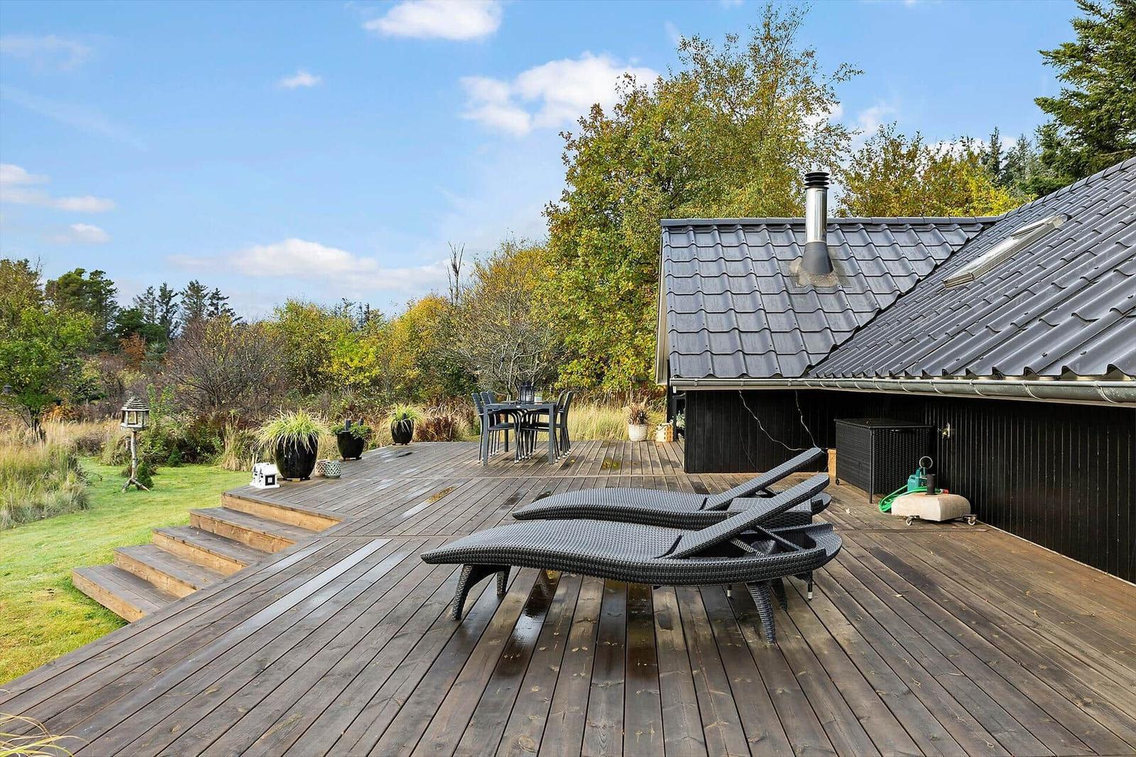 Wooden terrace with loungers, table, and chairs. Background: forest and house with roof.