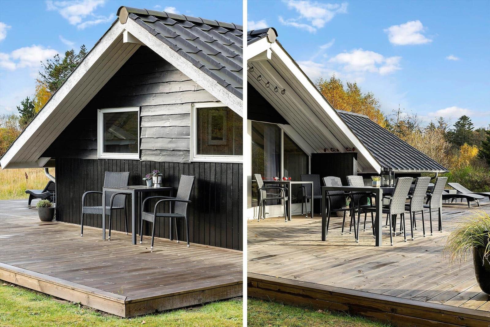 Wooden terrace with table and chairs in front of a house with dark siding and roof.