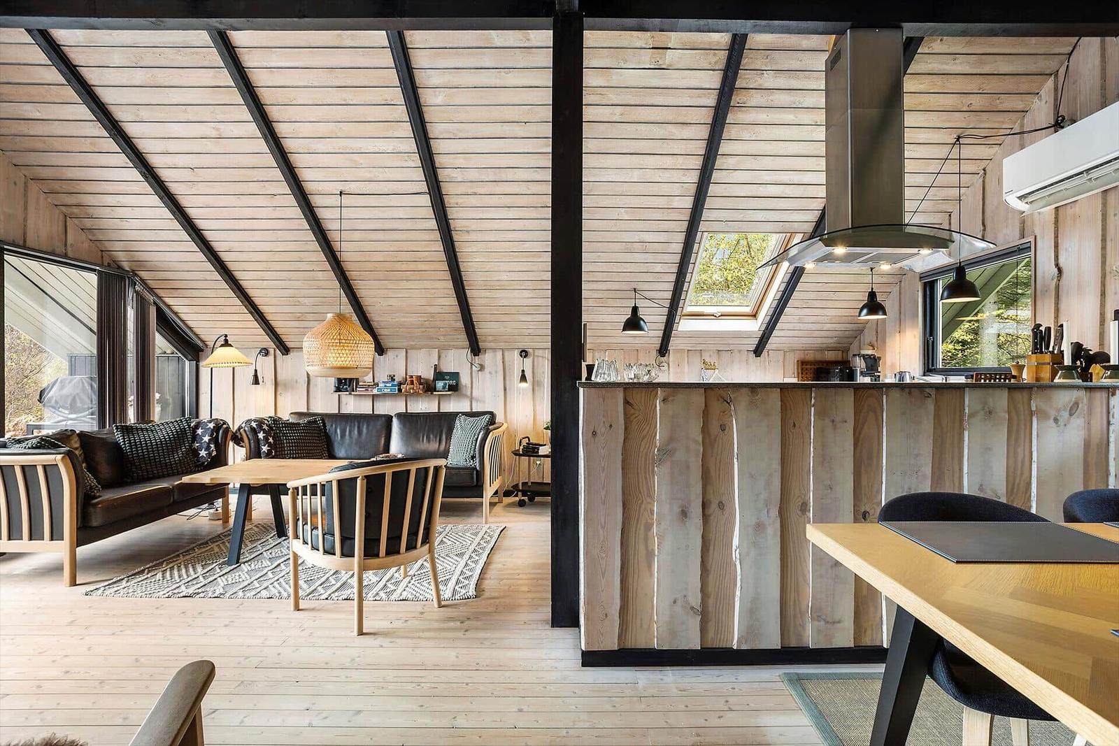 Living and kitchen area with wooden wall and ceiling. Dining table, sofa, and windows.