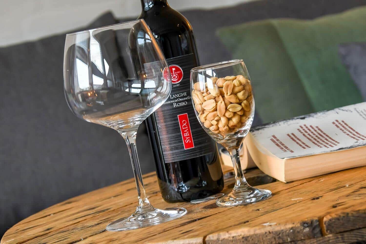 Ein Weinglas, eine Flasche Rotwein und eine Gläser mit Erdnüssen stehen auf einem Holztisch.