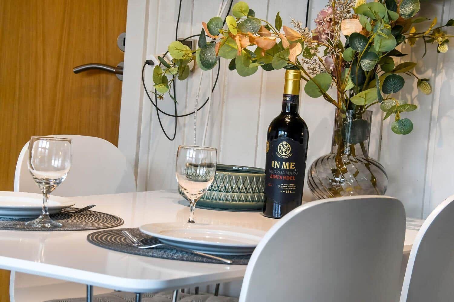 Esstisch mit Platz für zwei, Weinglas und Flasche, Blumenarrangement im Hintergrund.