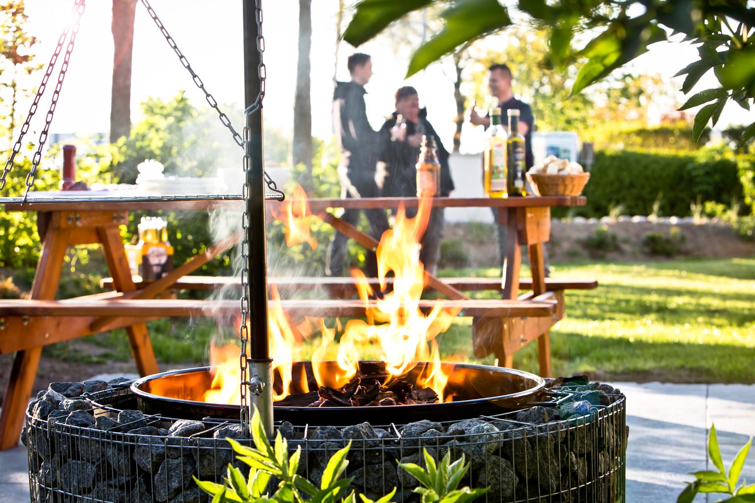 Ein brennender Feuerkorb mit Holz im Garten. Im Hintergrund steht ein Holztisch mit Personen.