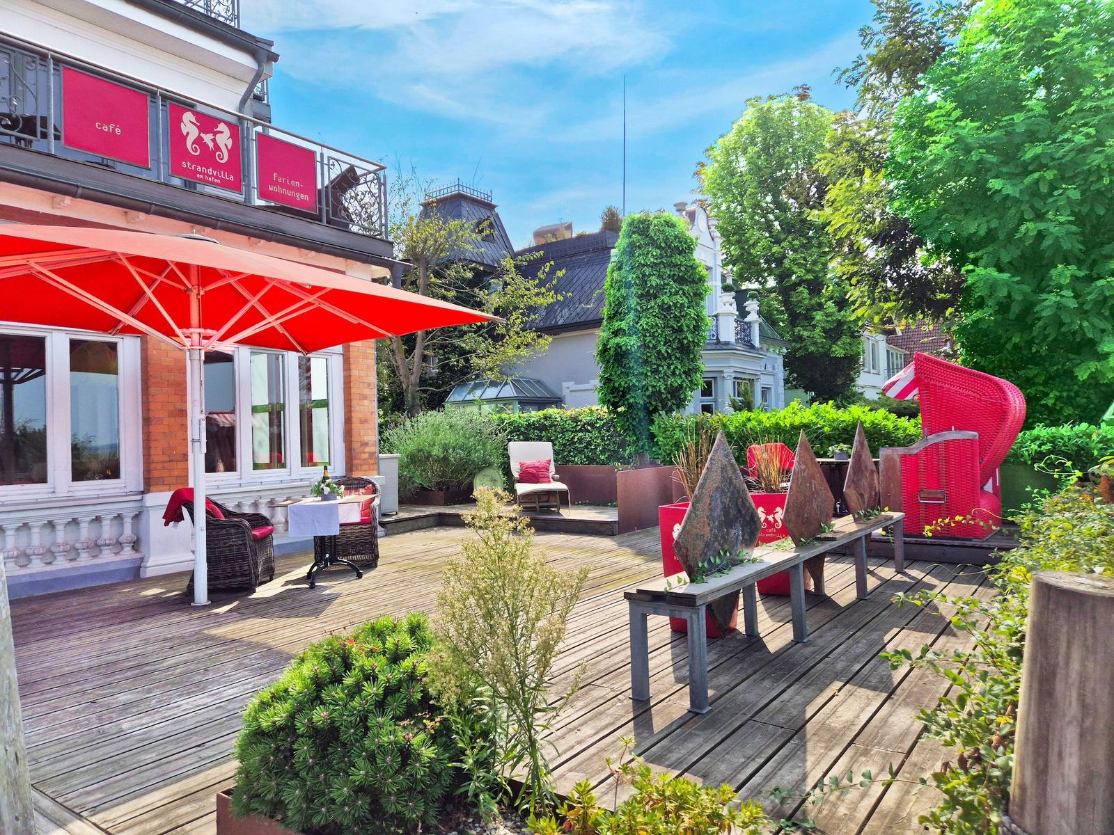Terrasse mit rotem Sonnenschirm und Gartenmöbeln. Holzdielen und Pflanzen umgeben die Loungezone.