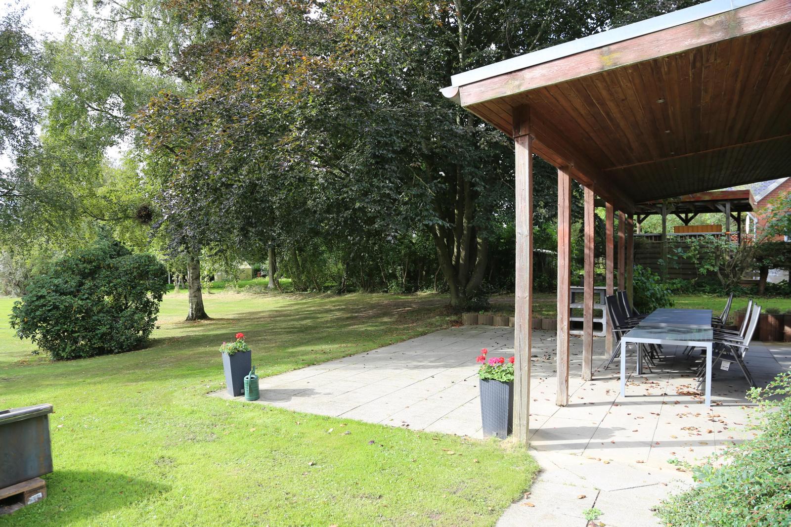 Patio with table and chairs under roof, surrounded by grass and trees.