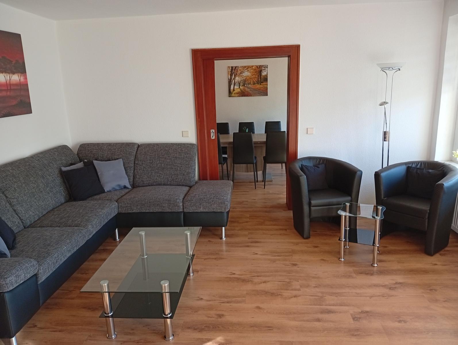 Wohnzimmer mit Sofa, zwei Sesseln und Glas-Tisch. Durchgang zur Essnische.