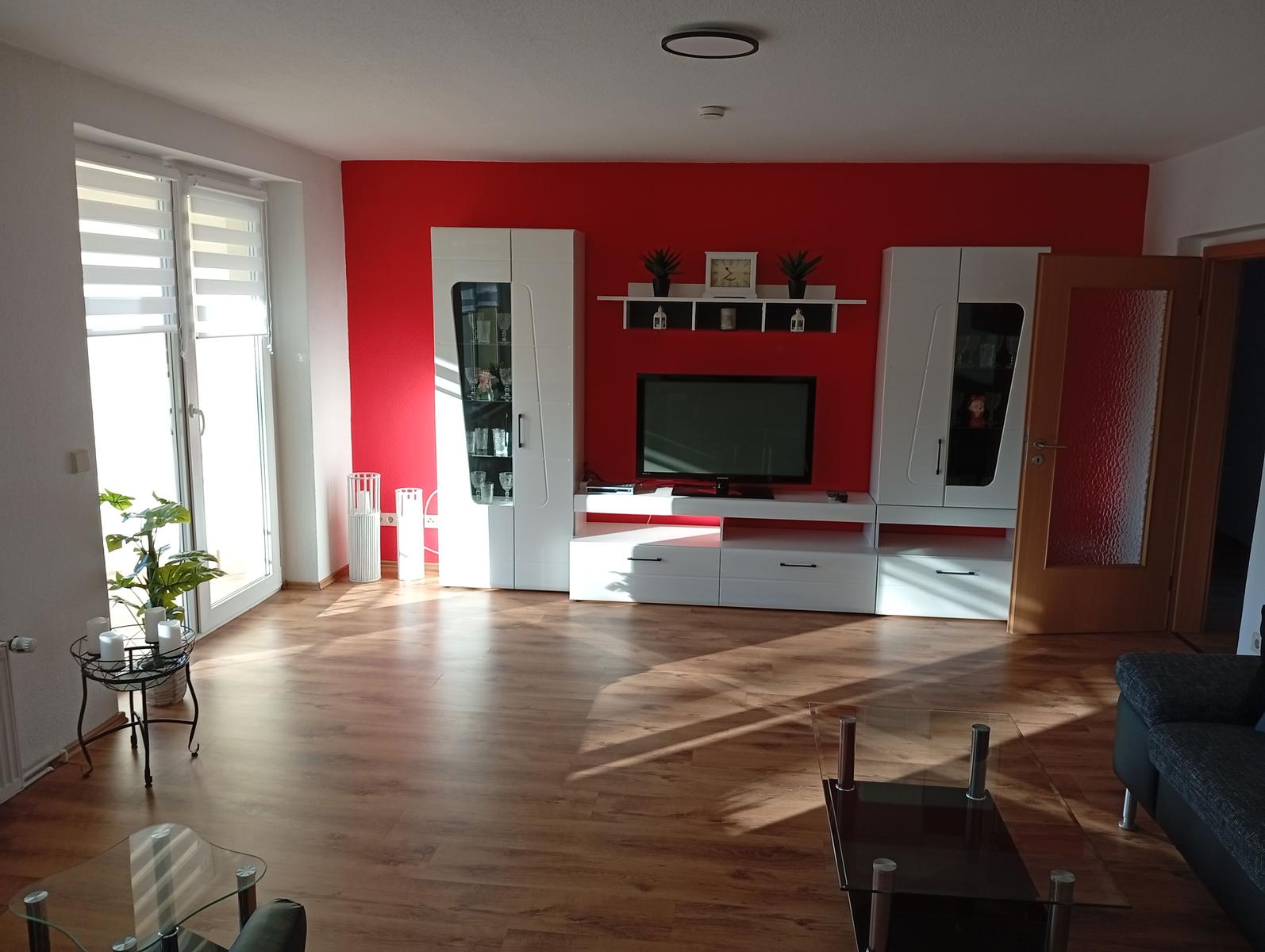 Wohnzimmer mit roter Wand, weißen Schränken und TV. Holzboden und Glas-Tisch.