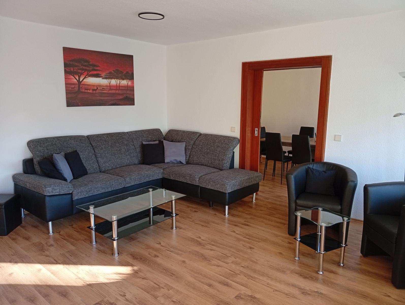 Wohnzimmer mit Sofa, Sesseln und Glas-Tisch. Durchgang zum Essbereich.