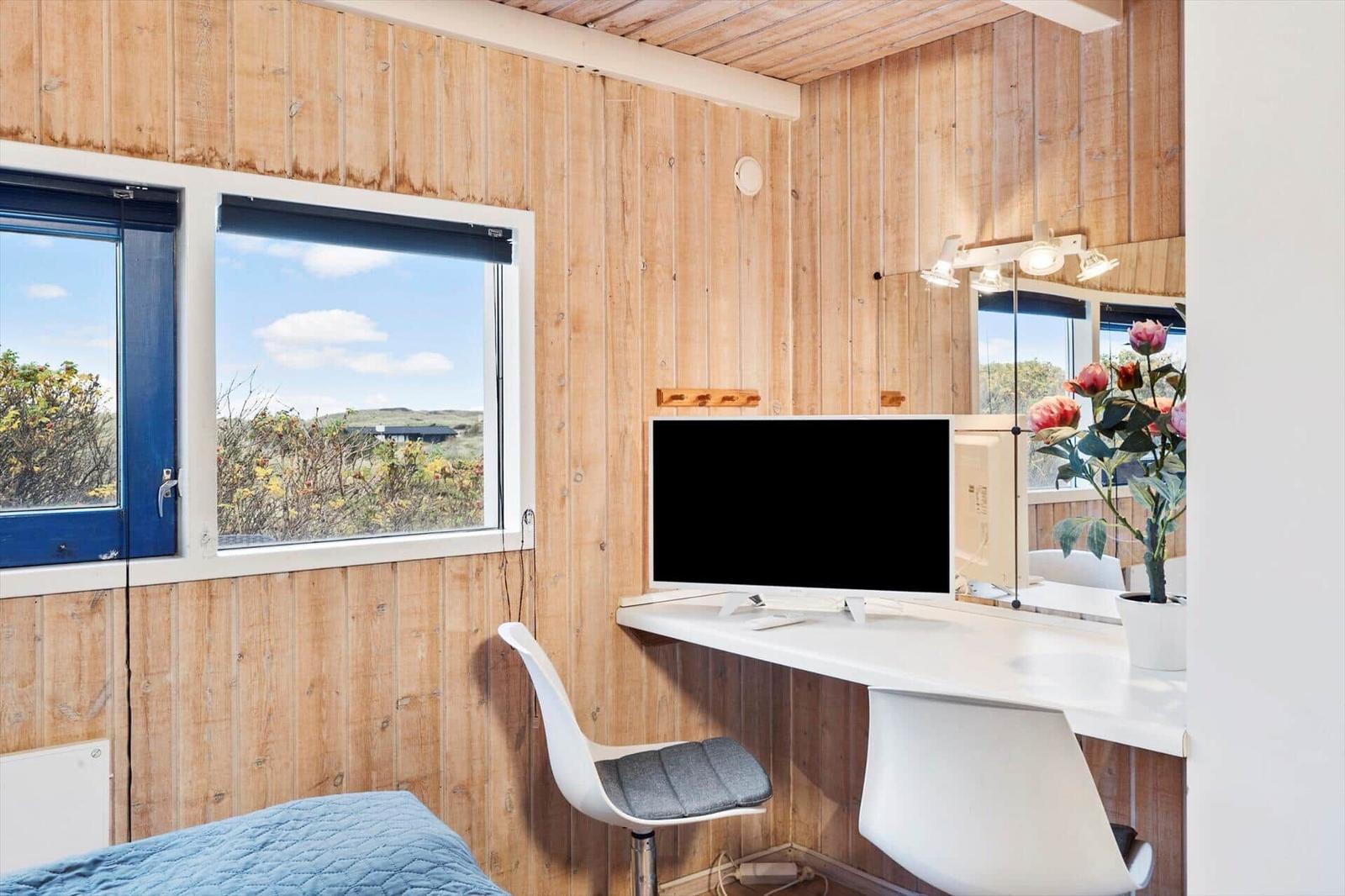 A room with wooden walls, desk, monitor, and bed. Window shows landscape.