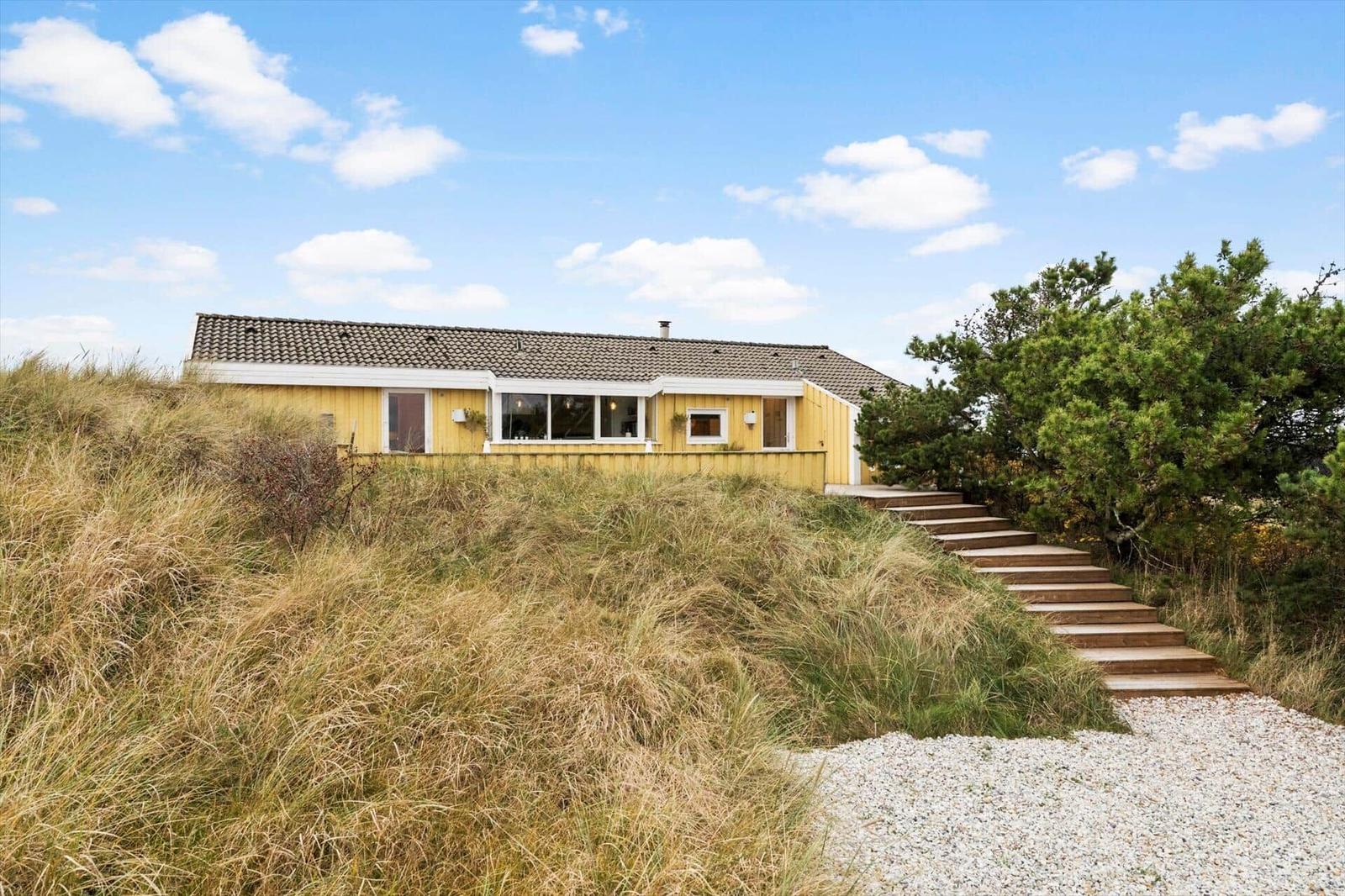 Yellow house with wooden steps and grassy slope under blue sky.