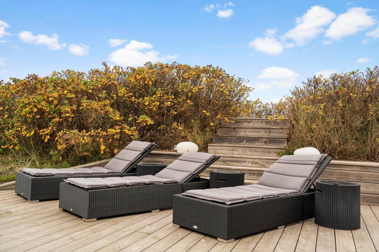 Three lounge chairs with cushions are on a wooden terrace in front of shrubs.
