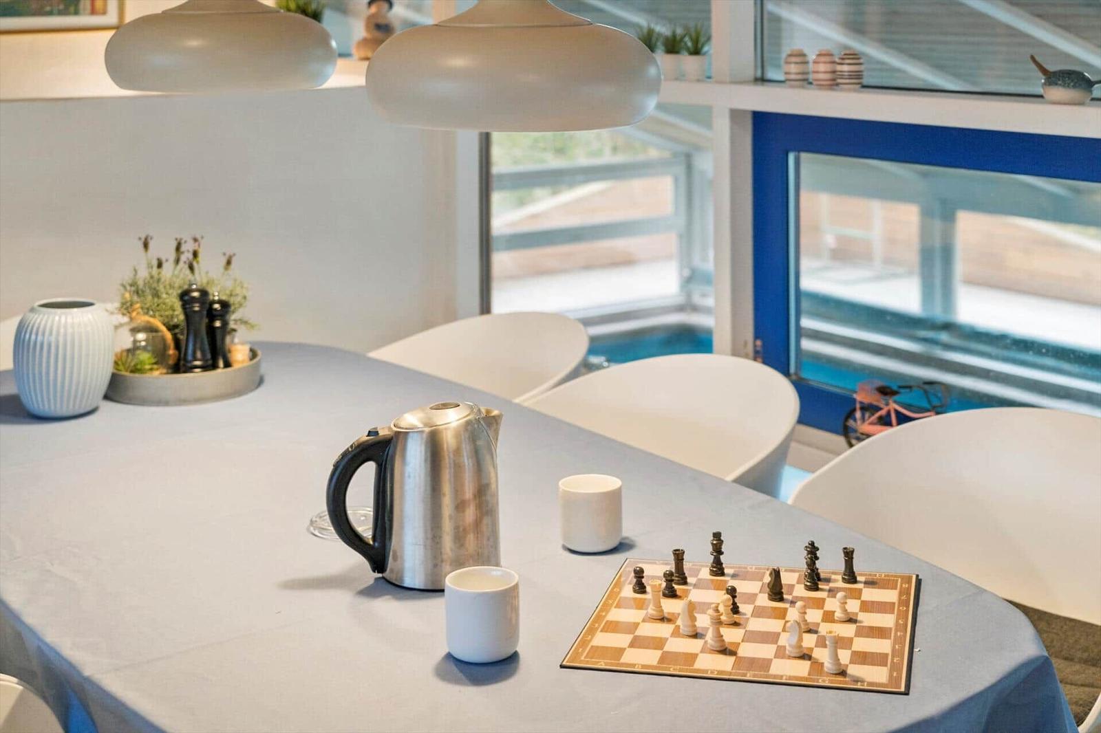Table with chessboard, kettle, and cups. White chairs and large windows.