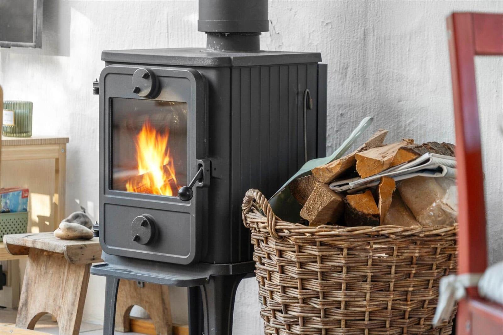 Schwarzer Holzofen mit brennendem Feuer und Korb mit Brennholz neben einem rustikalen Stuhl.