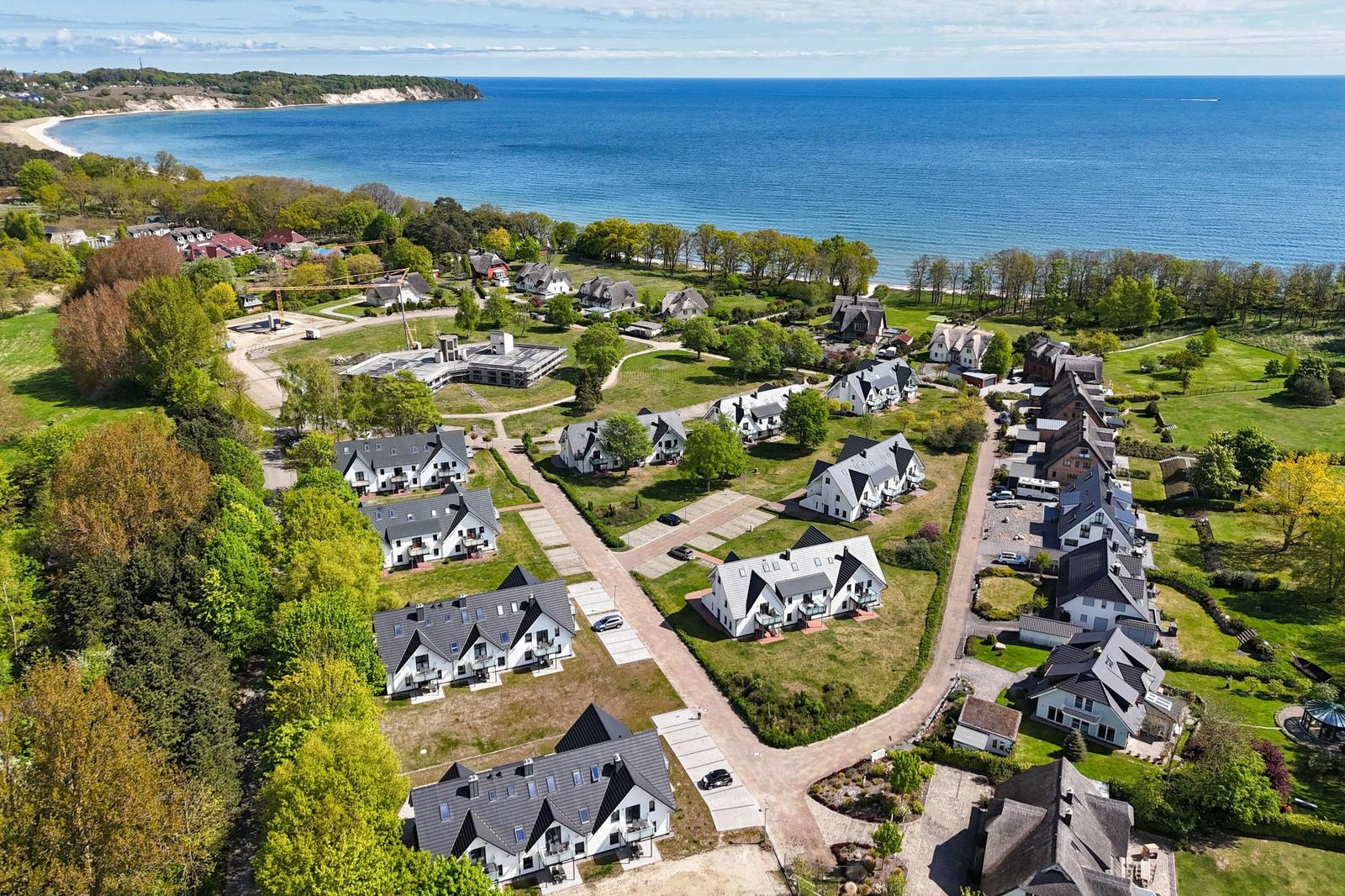 Aerial view of coastal residential area with houses, greenery, and sea in background.