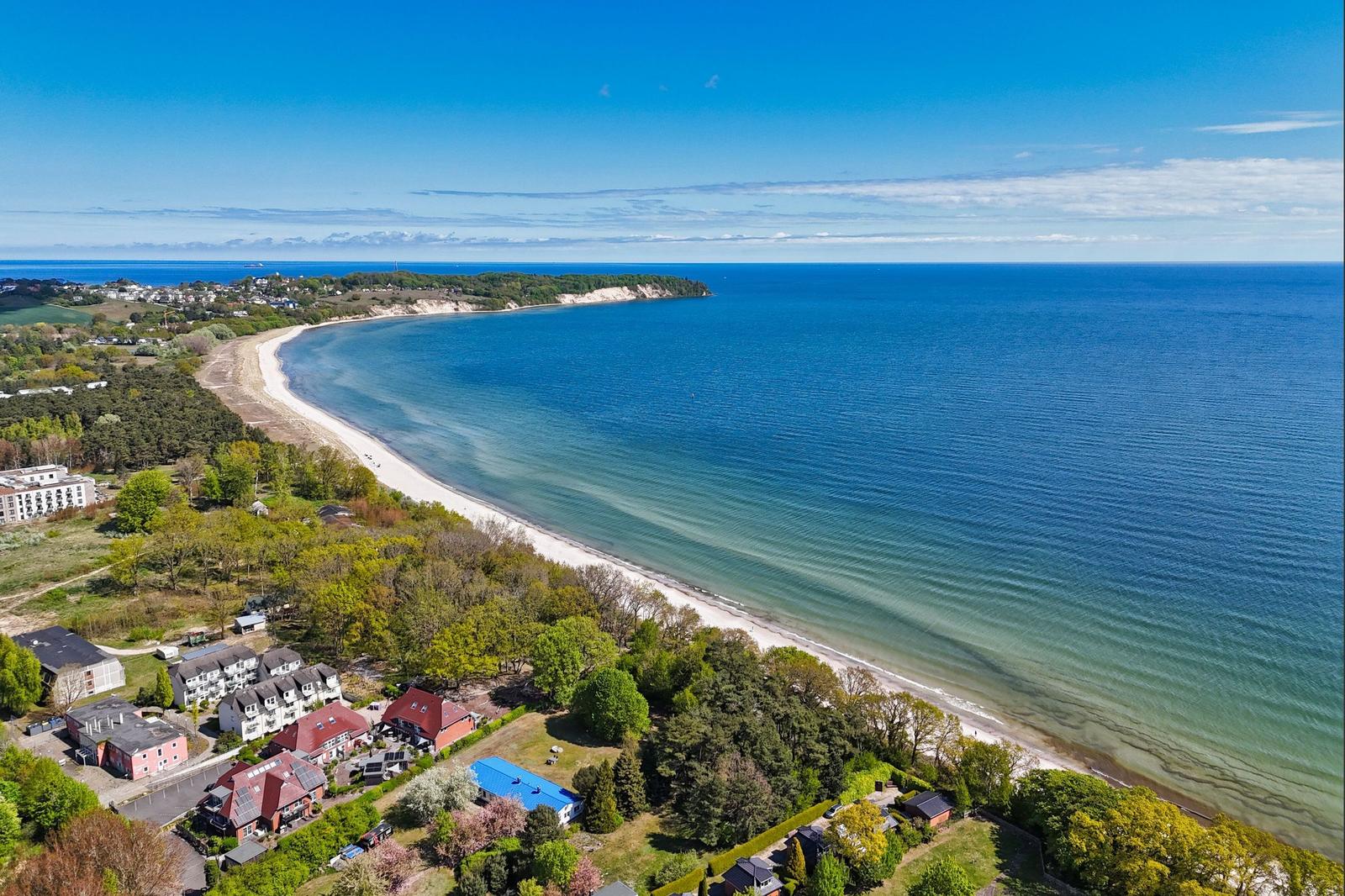 Aerial view of a beach with coastal houses and greenery under a clear blue sky.