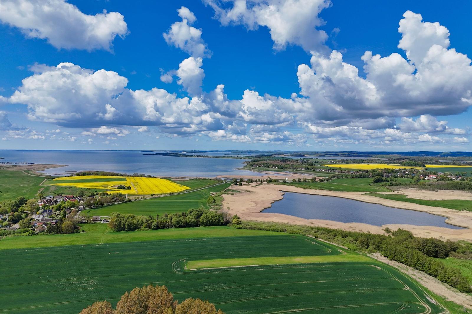 Landschaft mit Feldern, See und Küste unter blauem Himmel mit weißen Wolken.