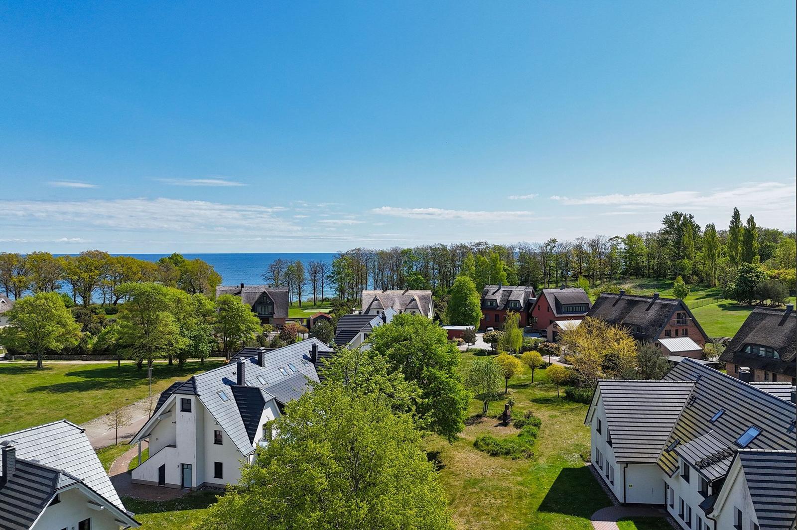 Häuser mit Dächern und Grünflächen, dahinter Meer und Bäume unter blauem Himmel.