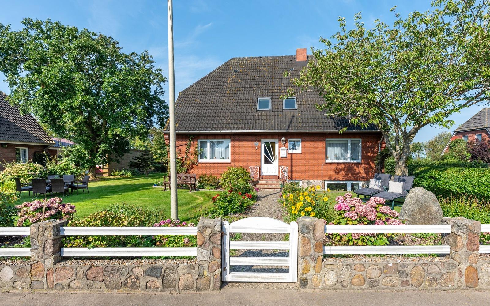 Rotes Haus mit Garten, Terrasse und Blumenbeeten. Weiße Toranlage und Holzbank.