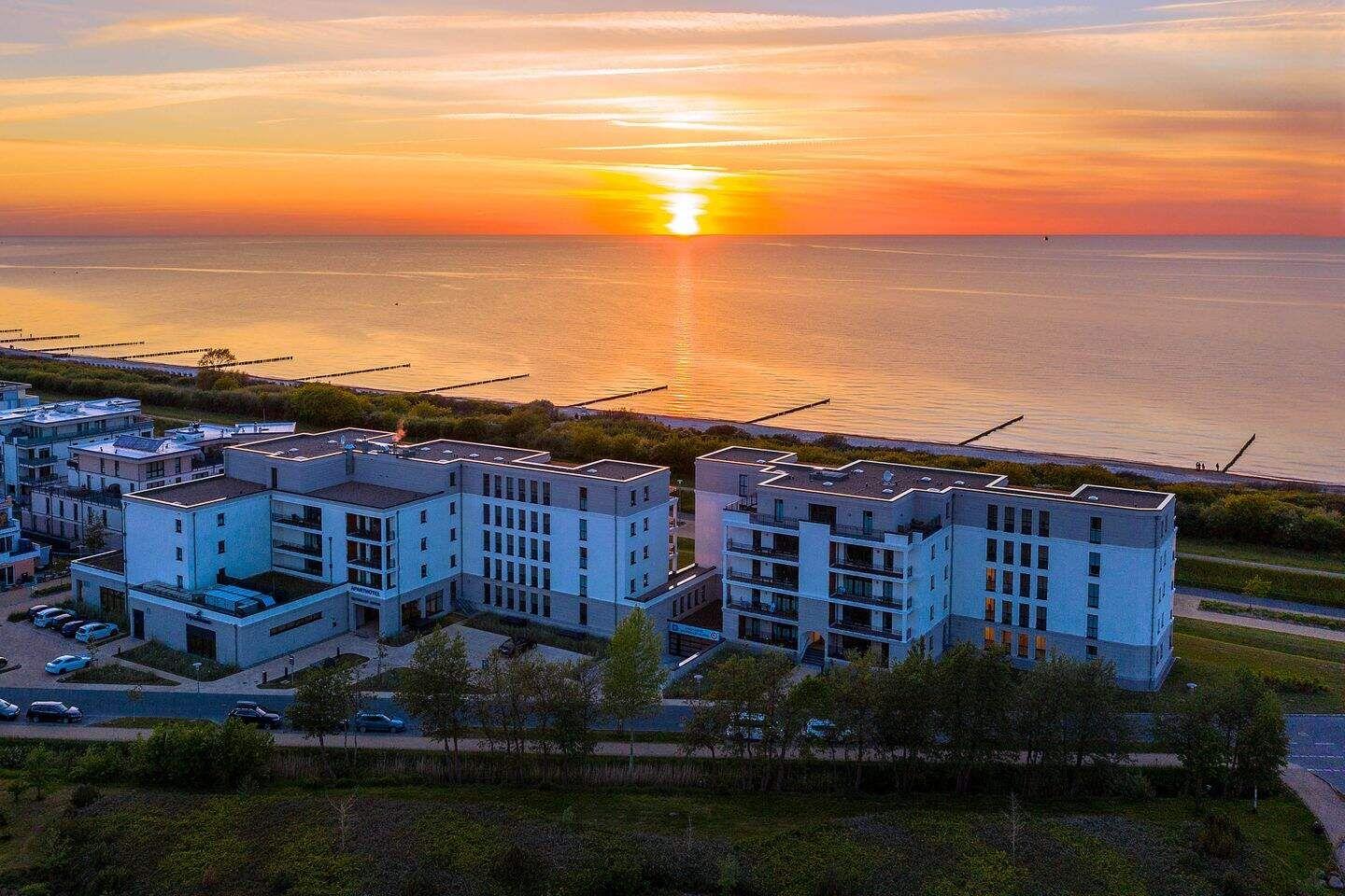 Modernes Gebäudekomplex am Meer bei Sonnenuntergang mit Blick auf ruhige Wasseroberfläche.