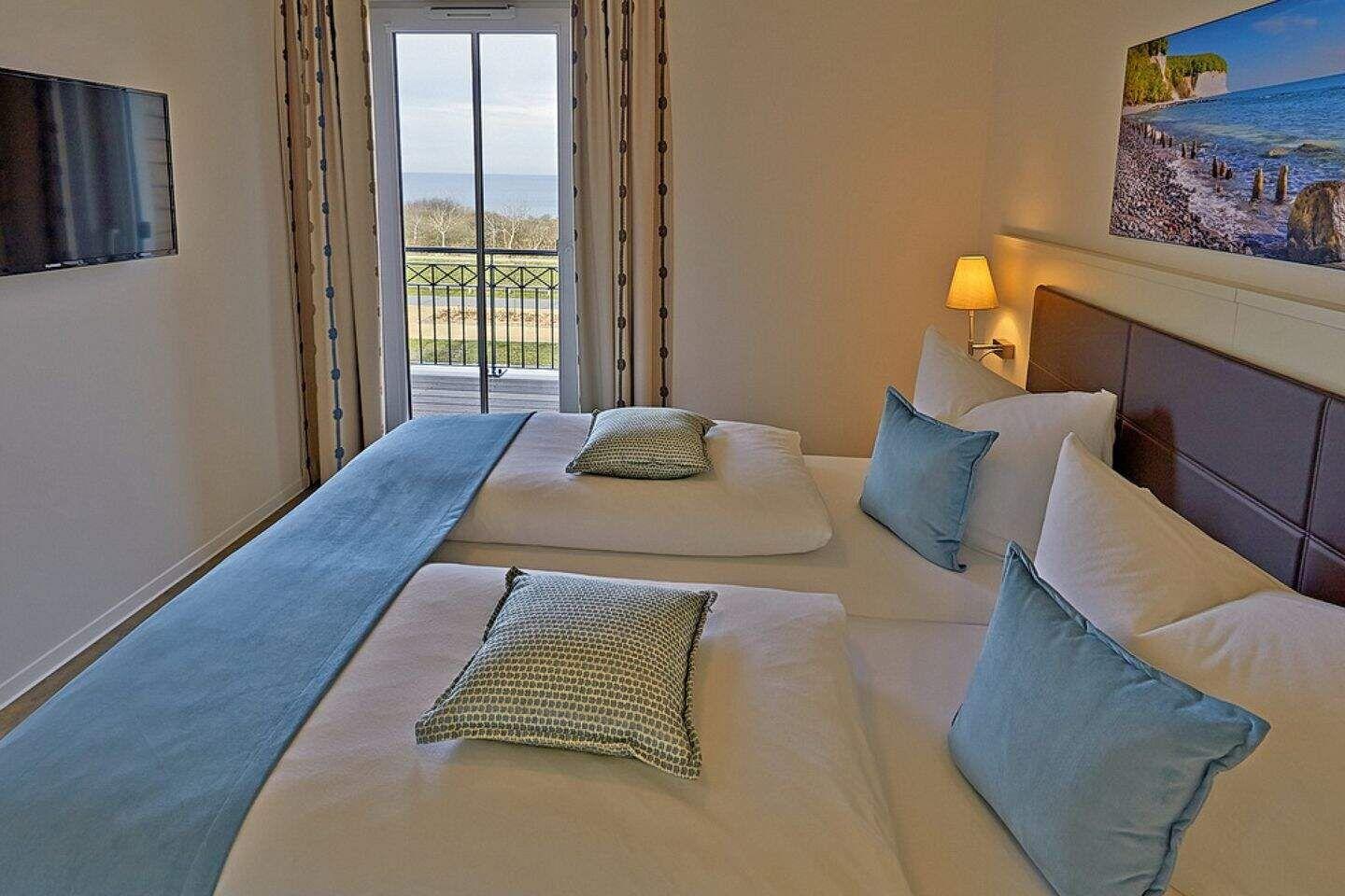Zwei Betten mit Kissen und Decke. Fenster zum Balkon mit Meerblick. Wandbild und Fernseher.
