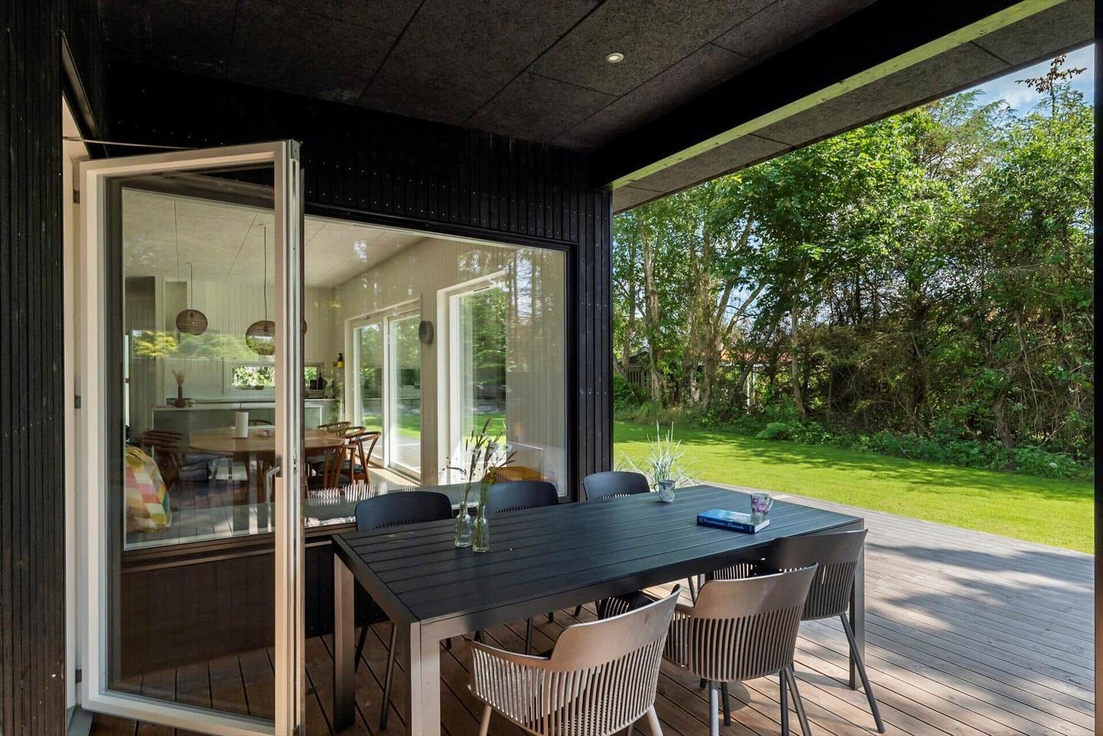Terrasse mit Tisch und Stühlen, Blick auf grünen Garten und Wald. Tür führt ins Haus.