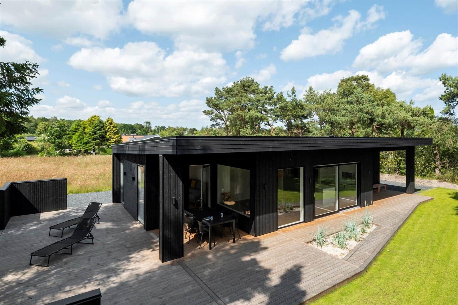 Moderne schwarze Hütte mit Terrasse, Liegen und Blick auf Wiese und Bäume.