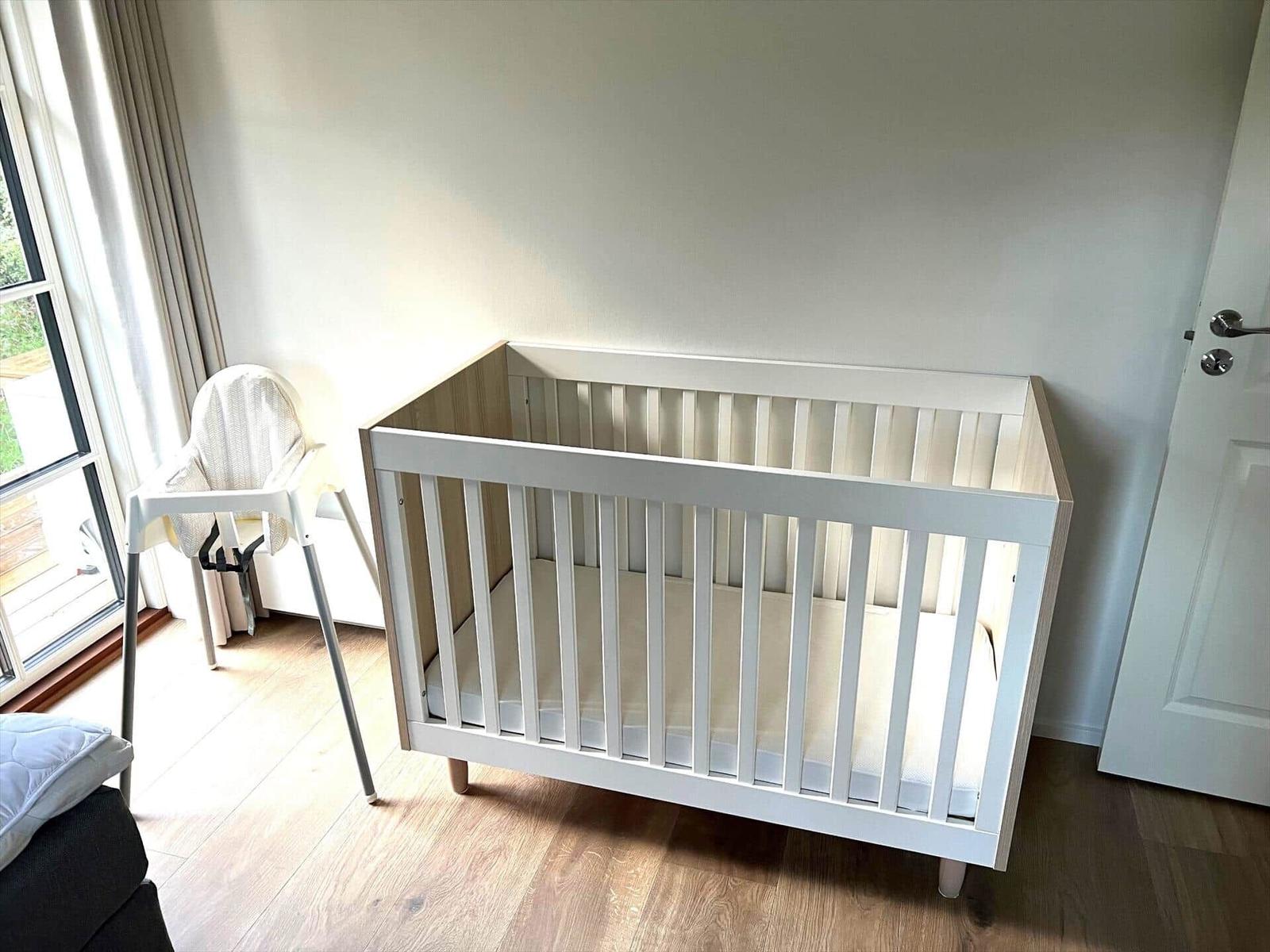 Kinderzimmer mit Hochstuhl und Babybett. Holzboden und Fenster mit Vorhang.