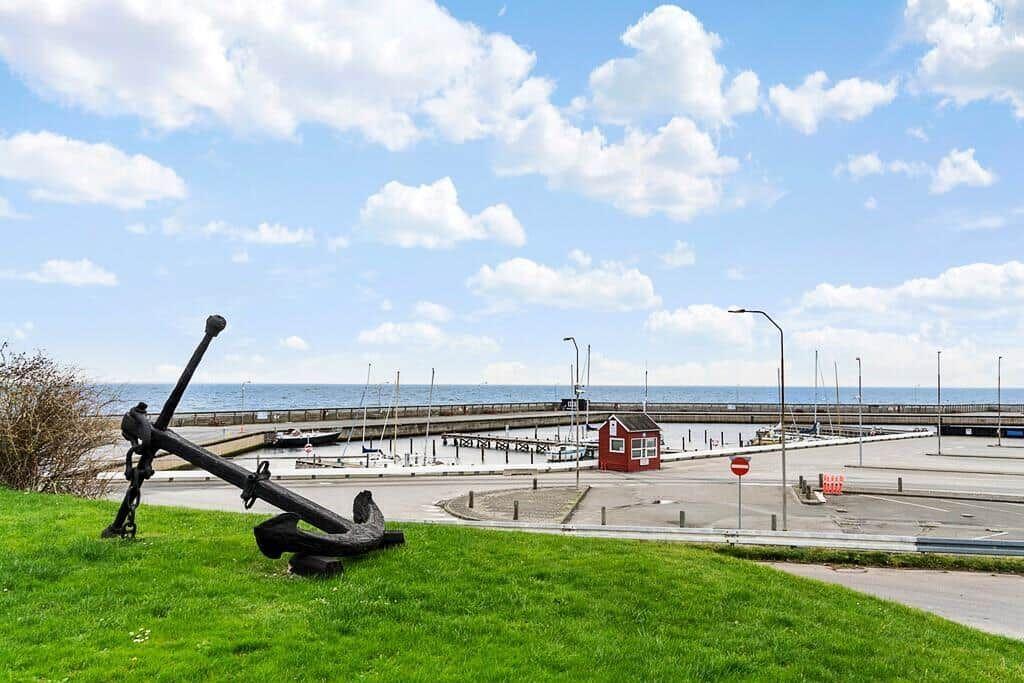 Großer schwarzer Anker auf grünem Rasen. Hintergrund: Hafen mit Booten und rotem Schuppen.