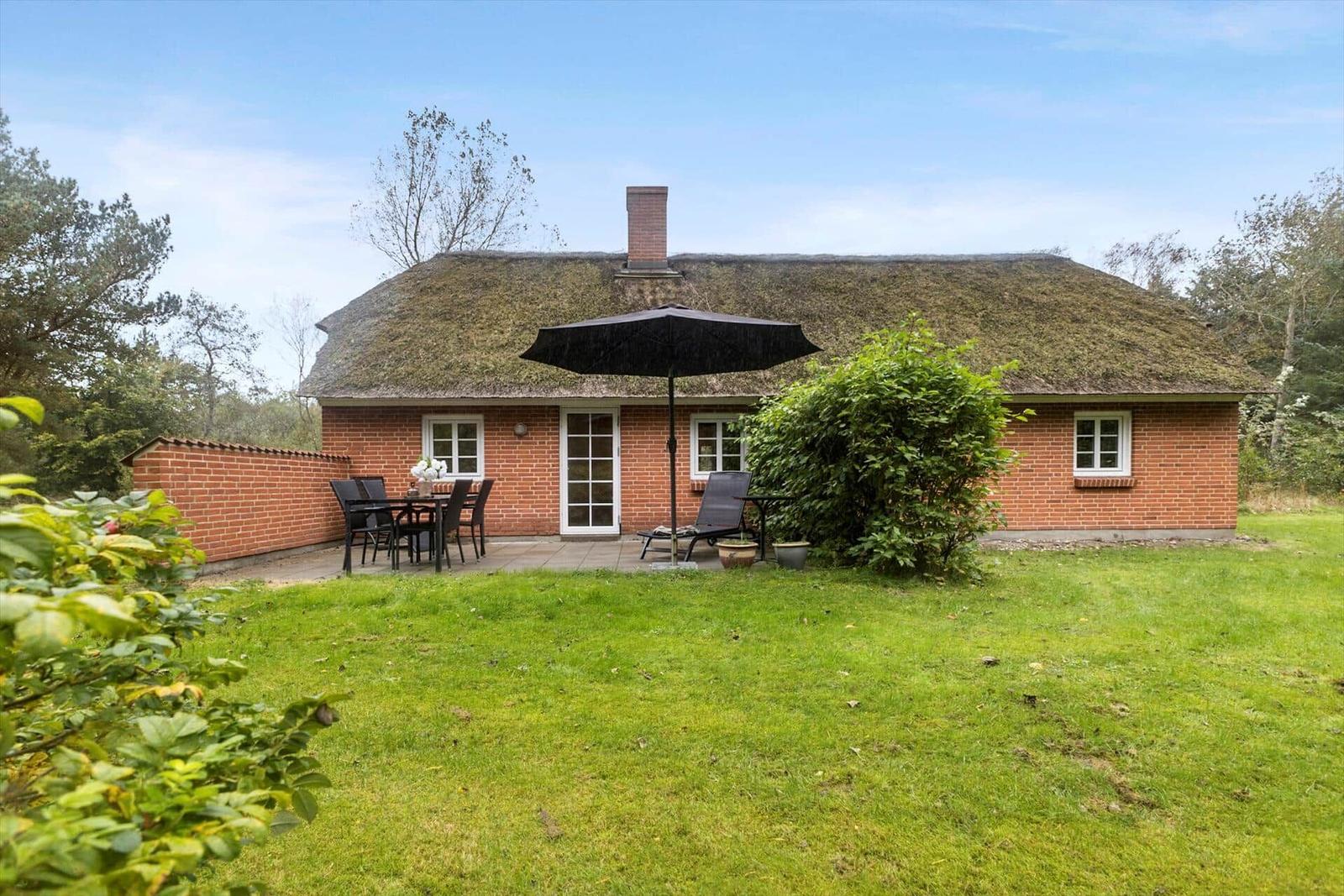 Rotes Backsteinhaus mit strohgedecktem Dach und Terrasse mit Tisch, Stühlen und Sonnenschirm.