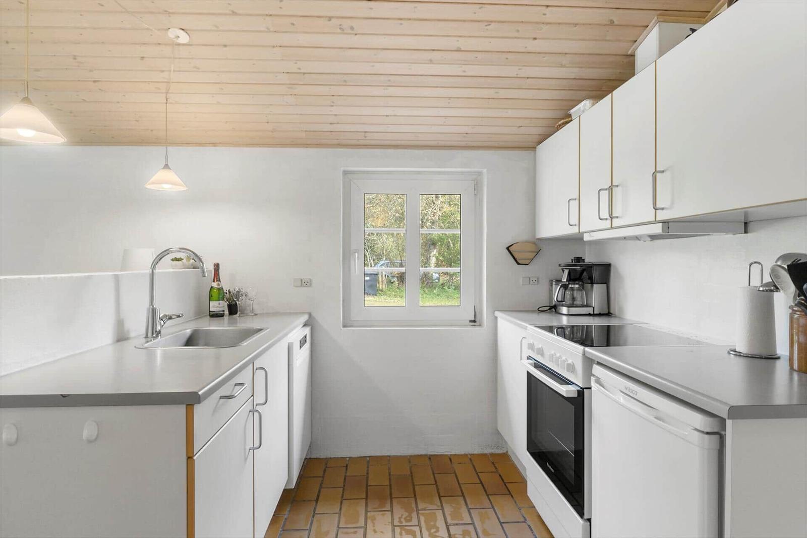 Küche mit weißen Schränken, Steinplatten und Holzdecke. Fenster zeigt grüne Landschaft.