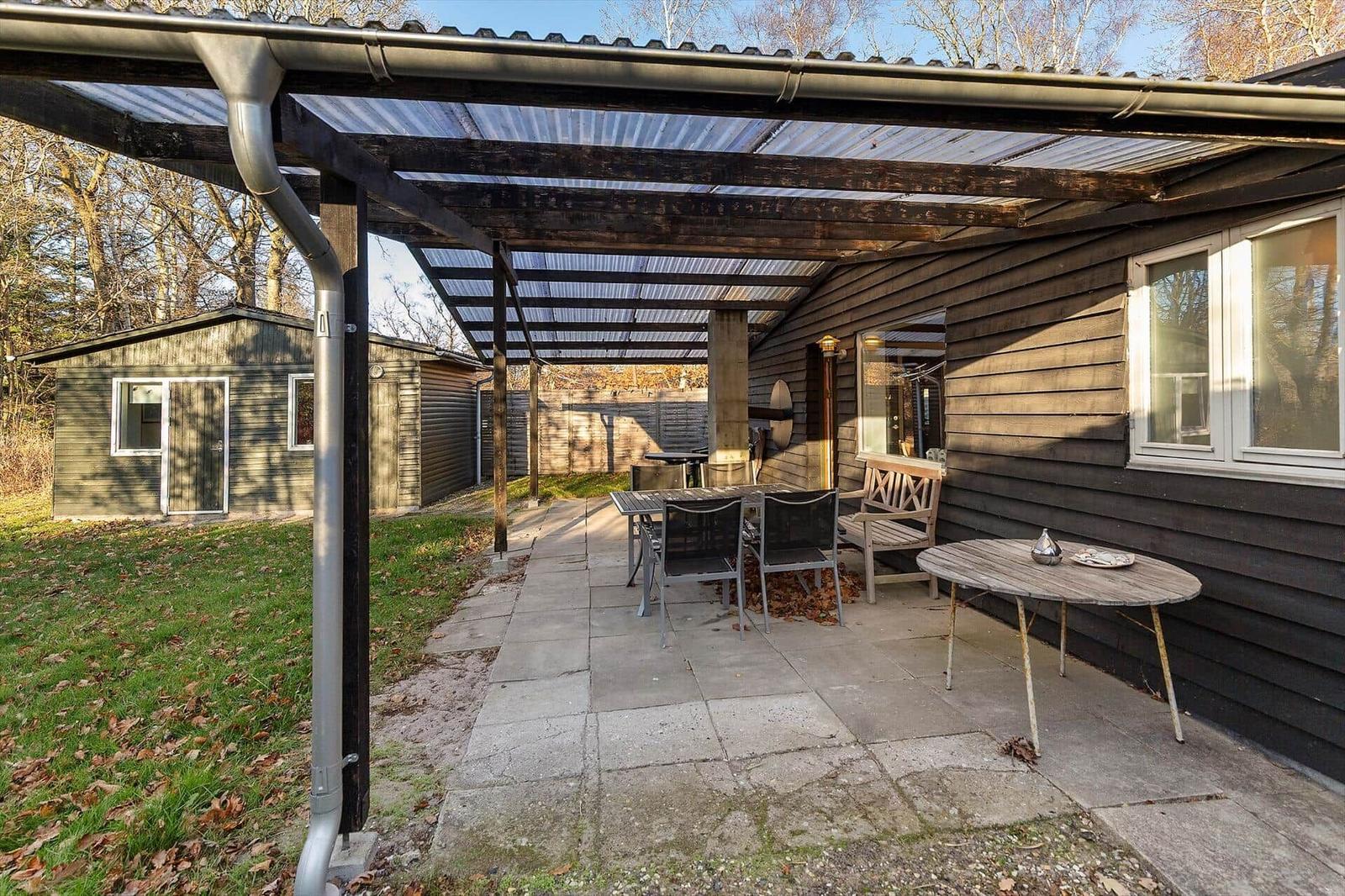 Terrace with pergola, tables and chairs beside wooden house. Lawn area and small outbuilding in background.