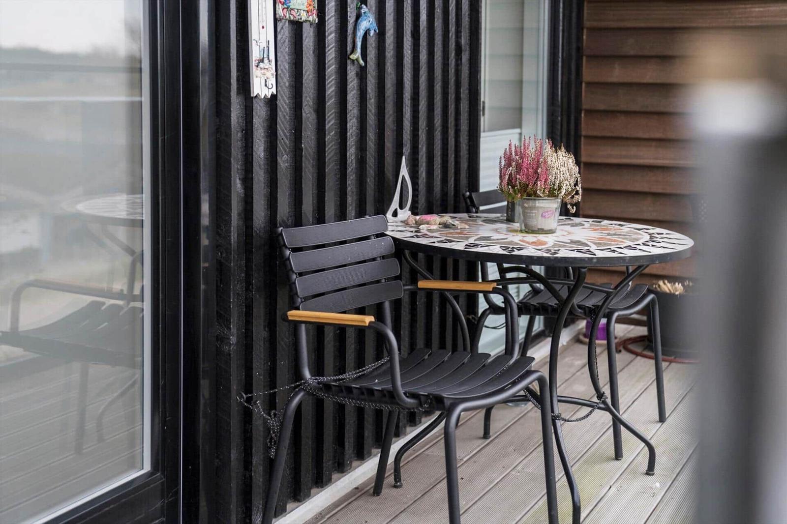 Terrasse mit Tisch und Stühlen, Blumen auf Tisch, schwarze Wand mit Dekoration.