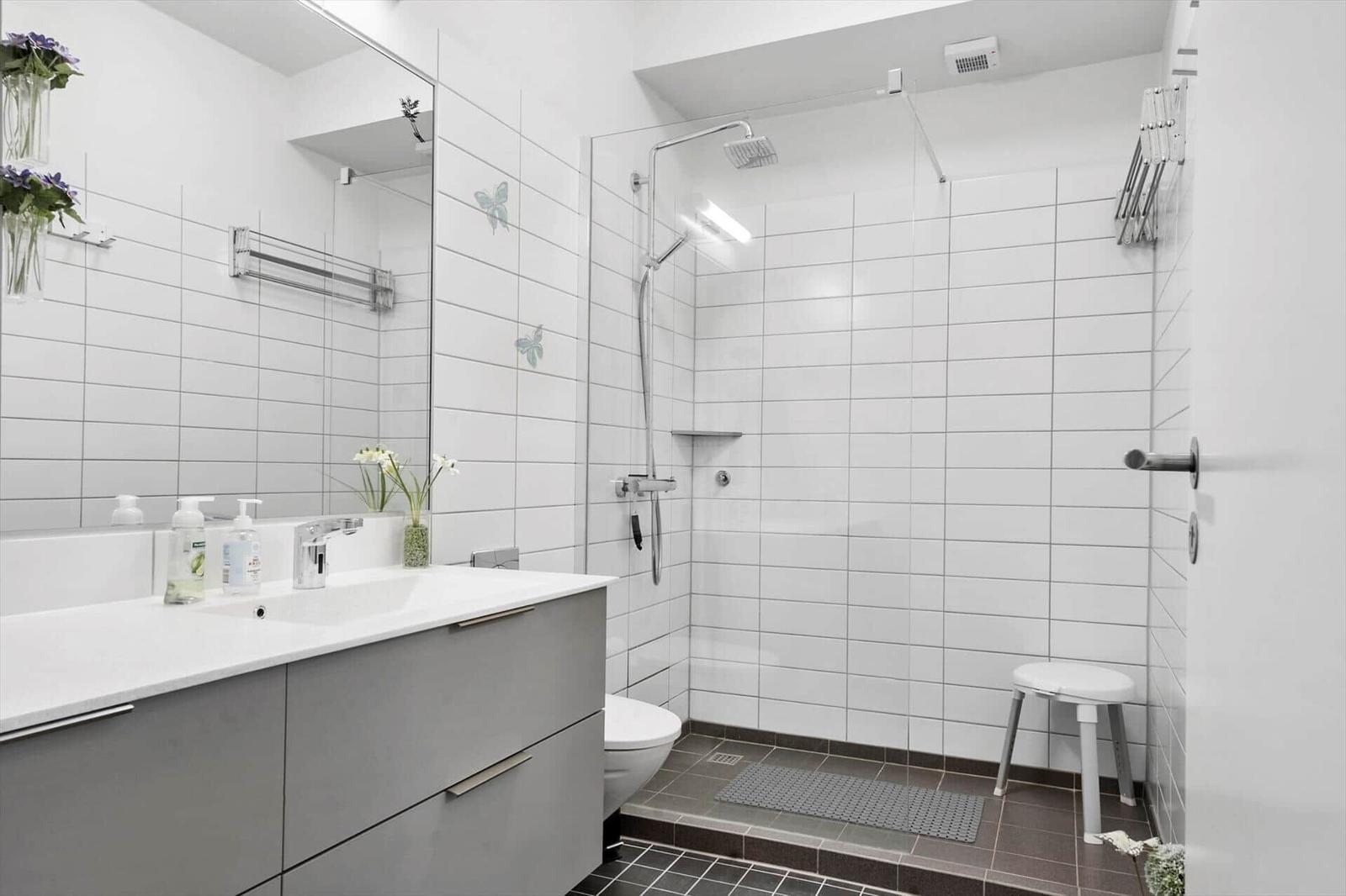 Badezimmer mit Dusche, Waschbecken und Toilette. Weiße Fliesen und graue Möbel.