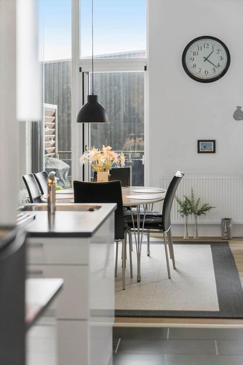 Essbereich mit Tisch, Stühlen und großem Fenster. Wanduhr und Pflanzen sind sichtbar.