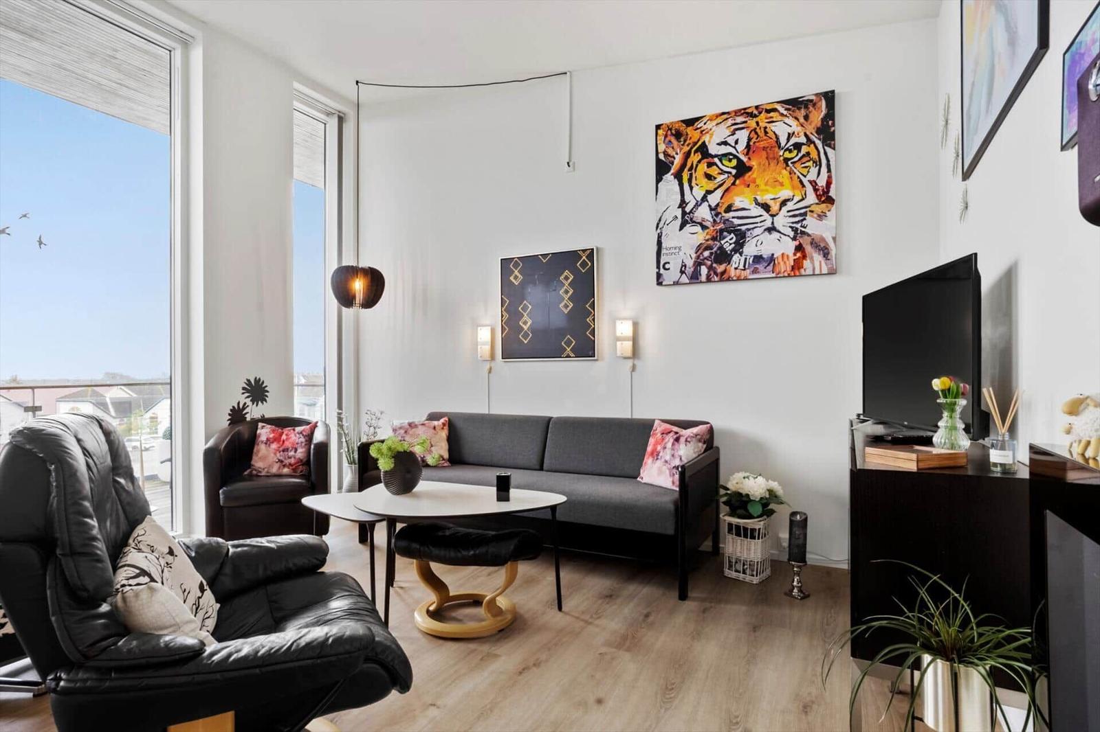 Wohnzimmer mit Sofa, Sessel und Fernseher. Große Fenster, Wandkunst und Pflanzen.
