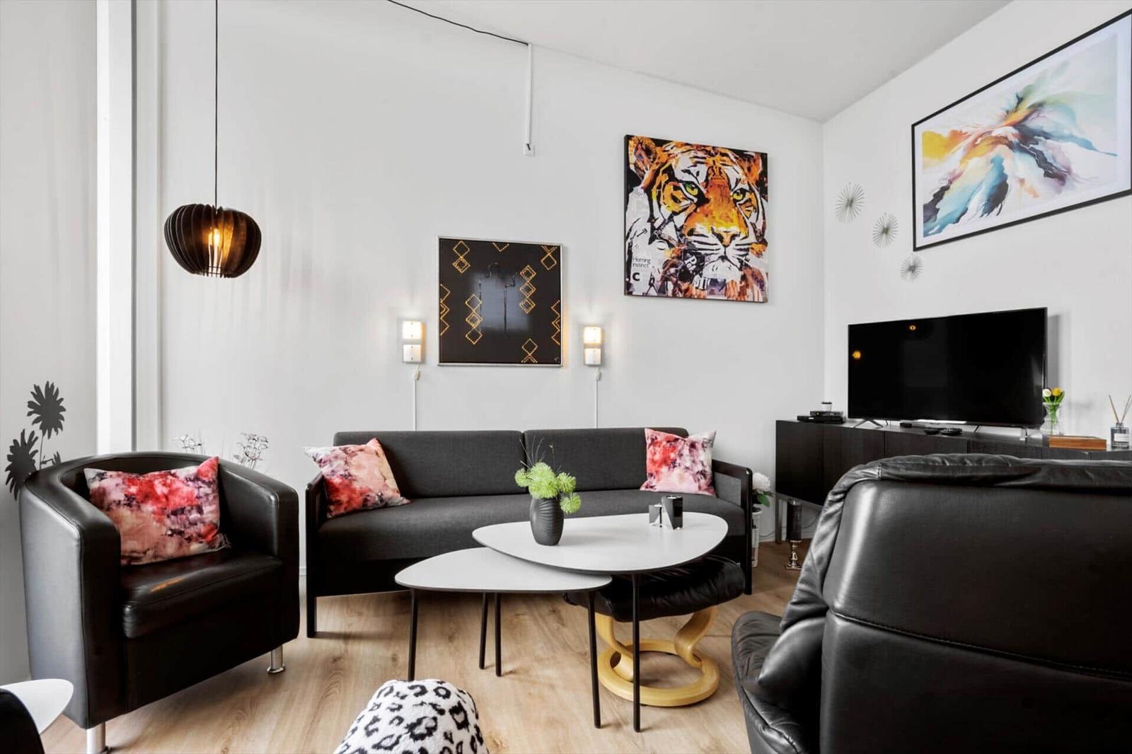 Wohnzimmer mit Sofa, Sessel und Couchtisch. Wandbilder und Fernseher sind sichtbar.