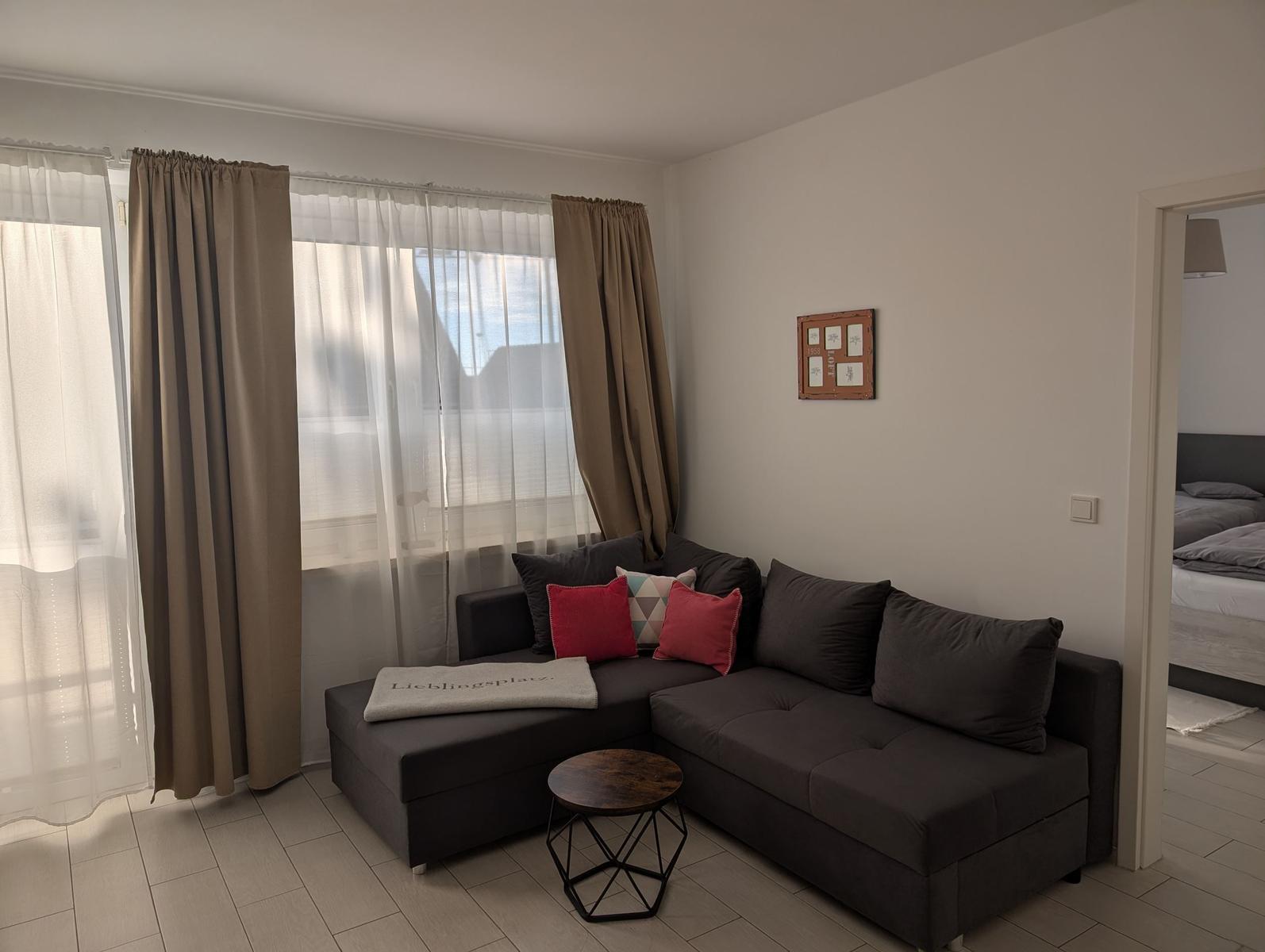 Wohnzimmer mit Ecksofa, Fenster und Blick in Schlafzimmer.