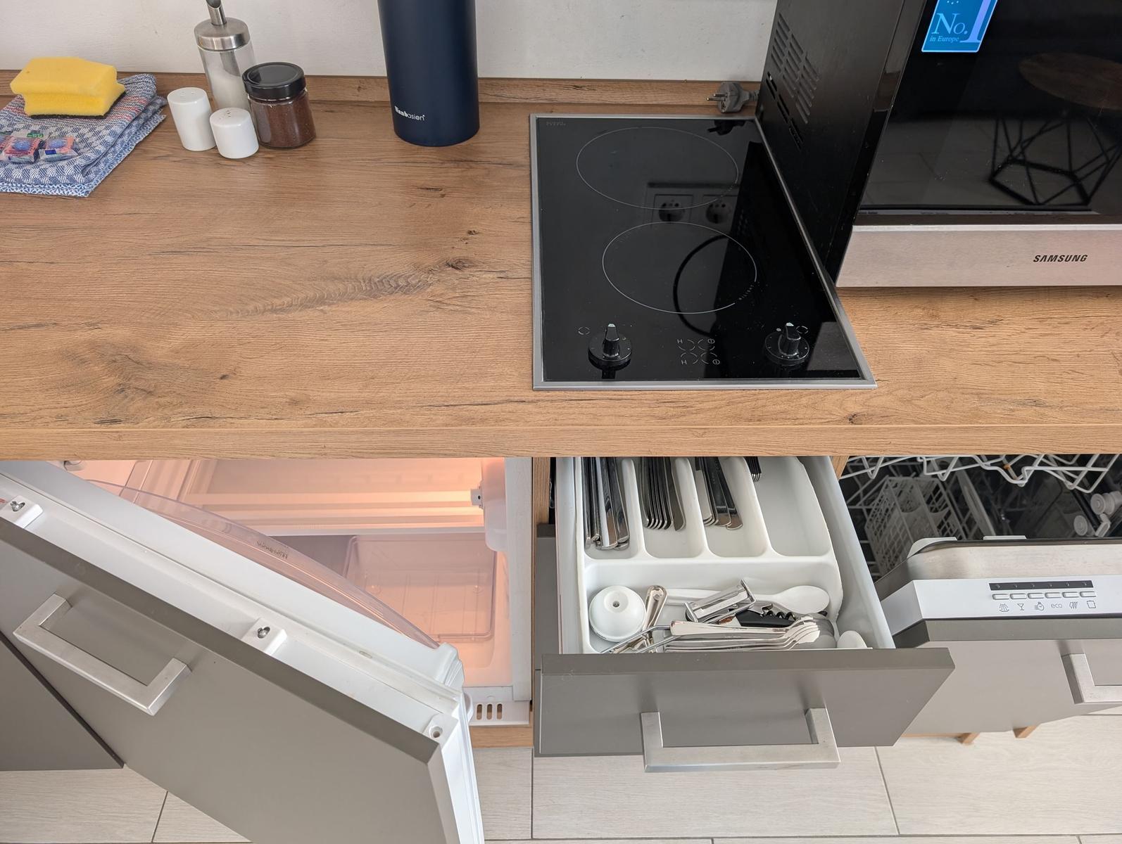Küche mit Herd, Spüle und Geschirrspüler. Besteckkasten und Kühlschrank sind sichtbar.