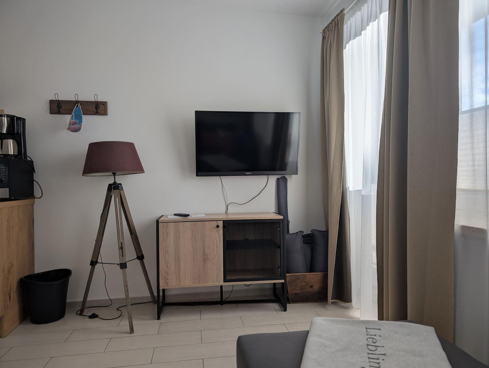 Wohnzimmer mit Fernseher, Stehlampe und Fenster mit Vorhängen.